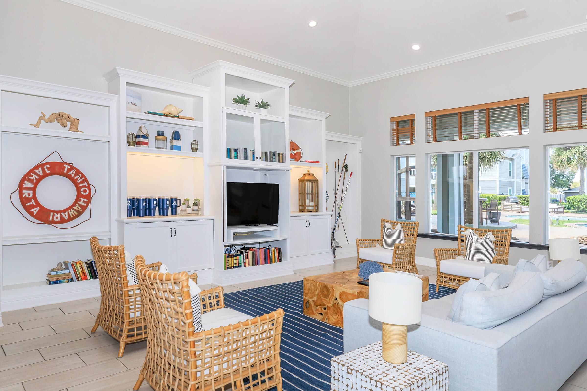 A bright and airy living room featuring a white sofa, woven chairs, and a decorative coffee table. The shelves are filled with books and decorative items. Large windows let in natural light, and a nautical-themed wall decor adds charm to the space. A blue and white striped rug anchors the area.
