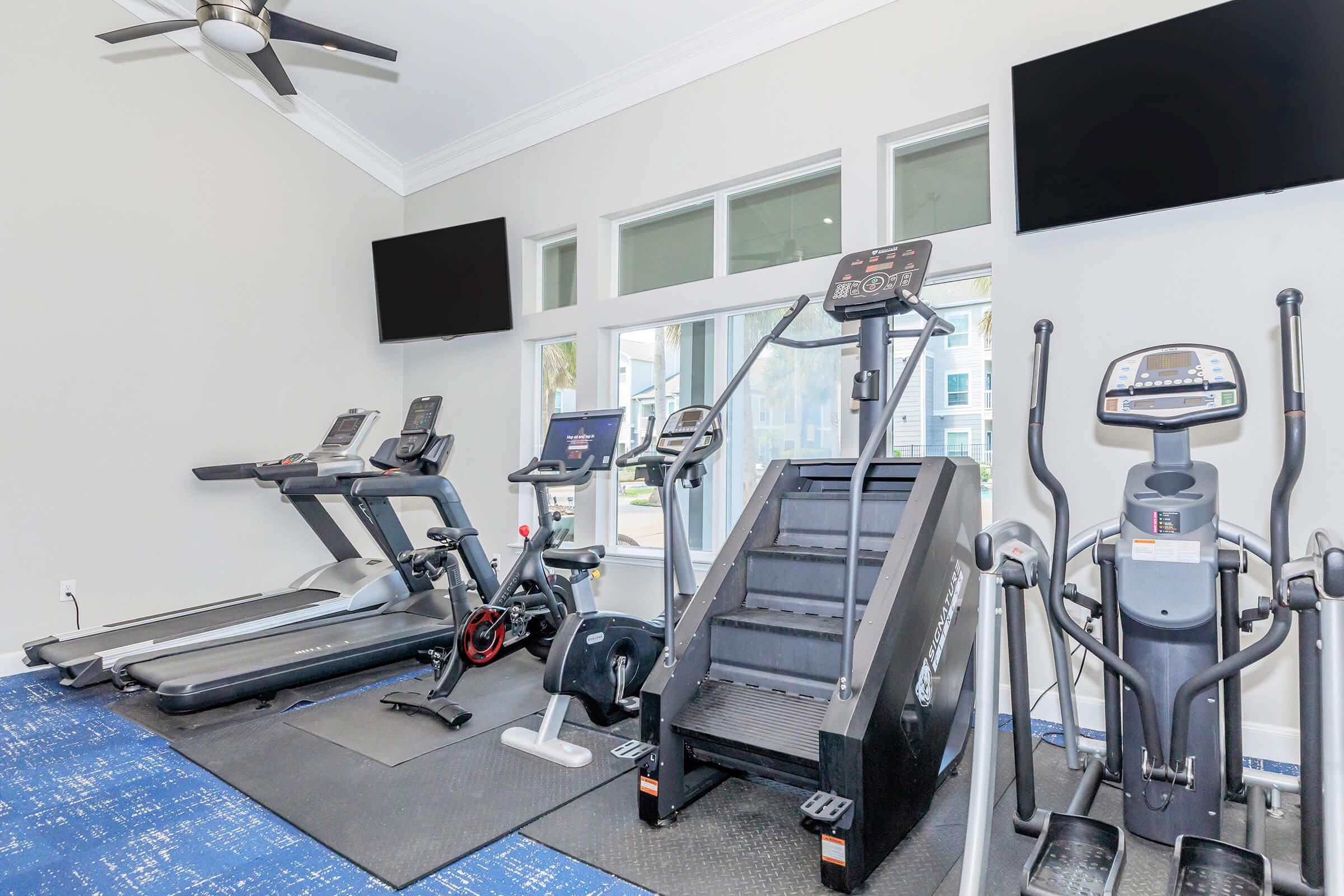 A modern fitness room featuring various exercise equipment, including treadmills, ellipticals, and a stair climber, with two wall-mounted televisions. Large windows allow natural light to illuminate the space, creating a bright and inviting atmosphere for workouts.