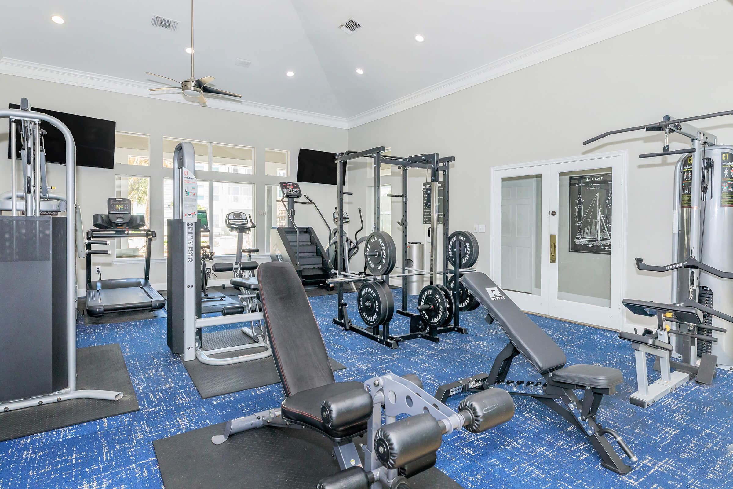 A modern gym interior featuring various fitness equipment, including weight machines, free weights, treadmills, and benches. The space is brightly lit with large windows, has a blue carpet, and includes two wall-mounted televisions for entertainment.