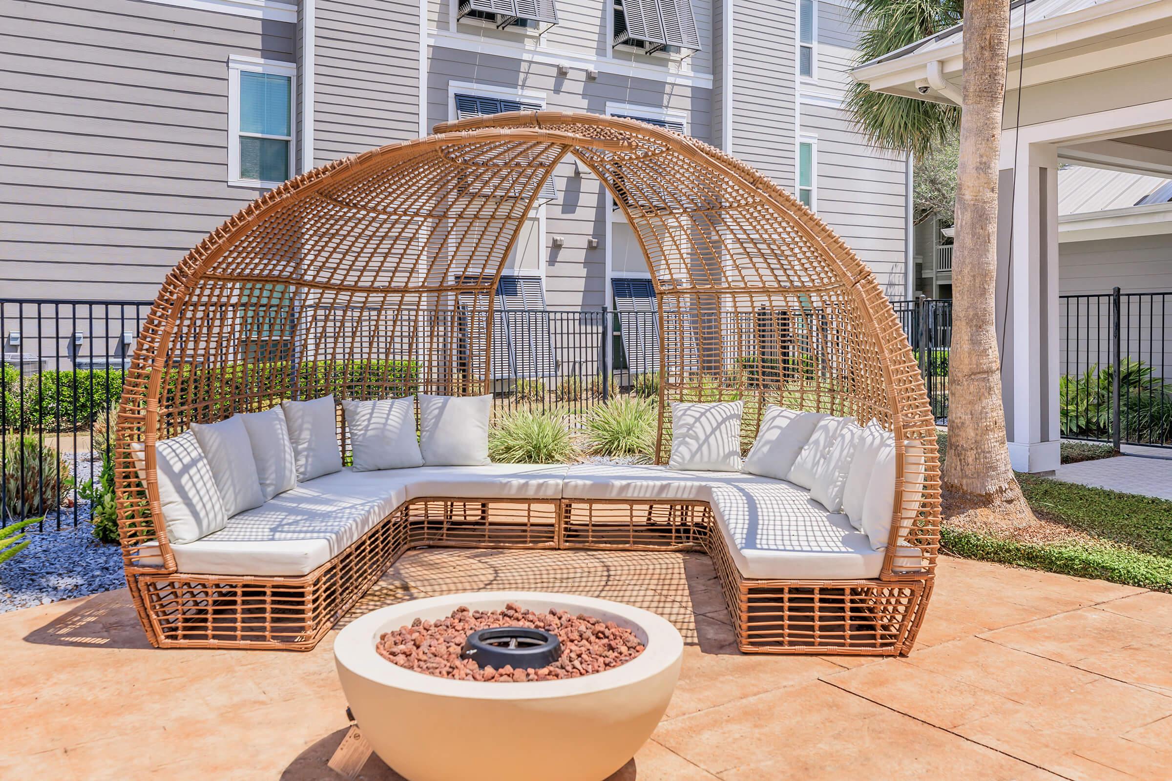 Stylish outdoor seating area featuring a curved, wicker-style canopy couch with white cushions, surrounded by greenery and a modern fire pit in a contemporary setting.
