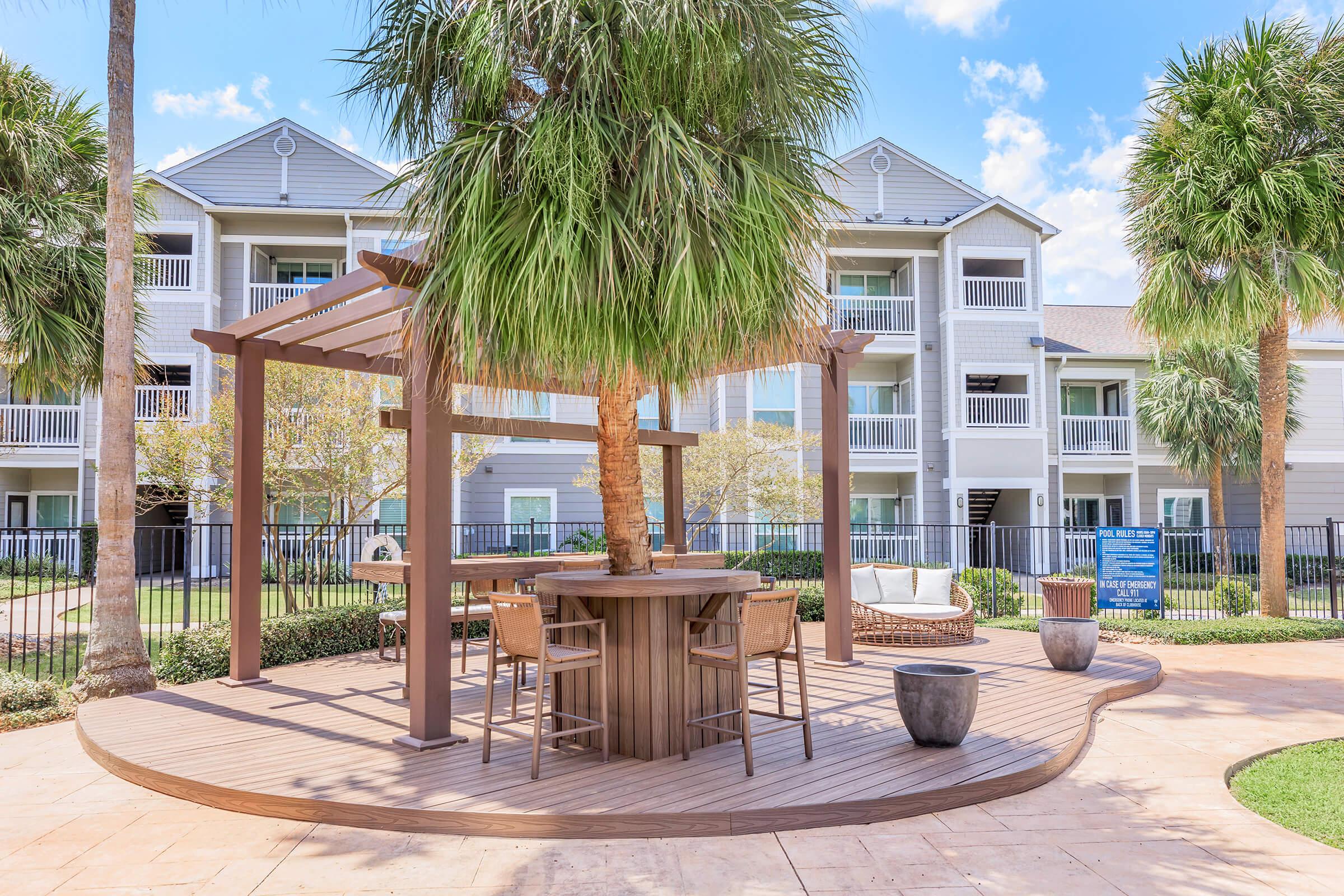 A wooden gazebo with bar-style seating surrounded by palm trees, set in a landscaped area with a well-maintained lawn. In the background, a multi-story building with balconies is visible under a blue sky with scattered clouds.