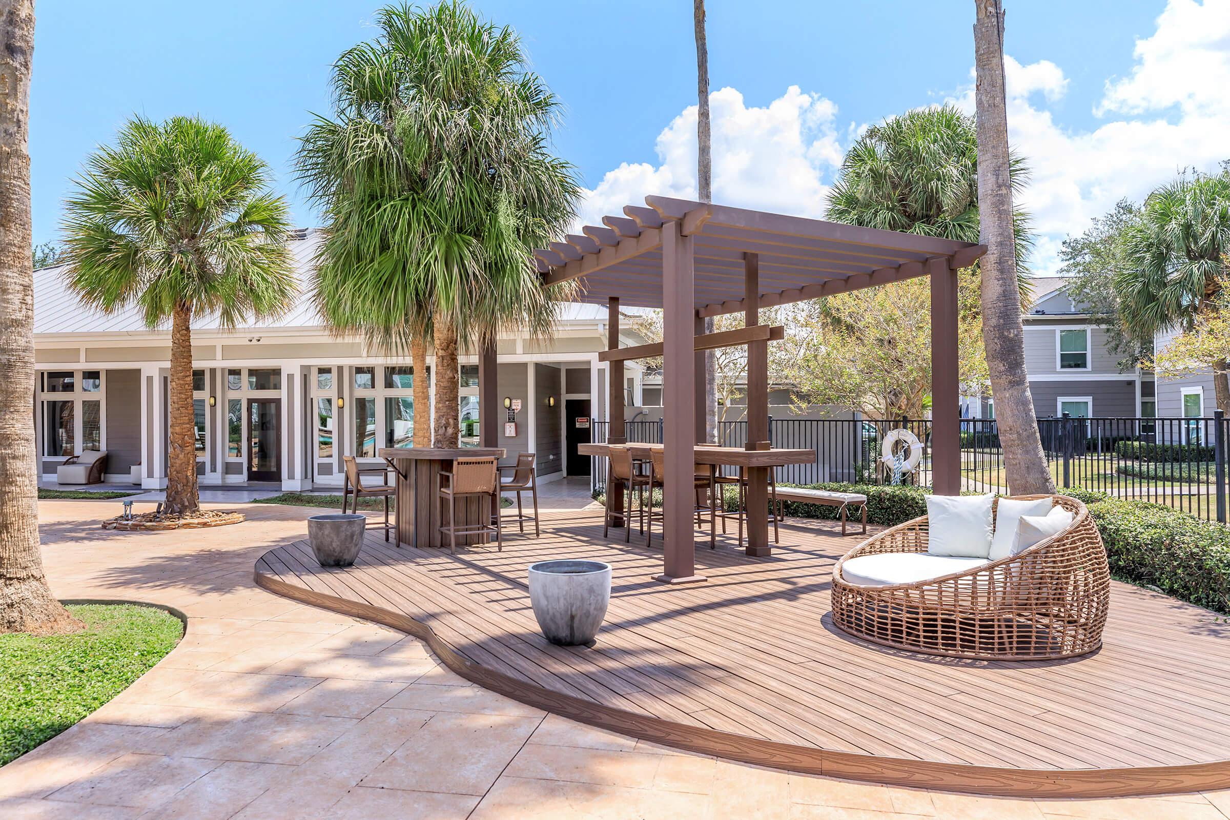 Outdoor patio area featuring a wooden pergola with seating, surrounded by palm trees. The space includes a round white chair and a small table, with a backdrop of a modern building and a pool area visible in the distance. The scene is bright and well-lit, suggesting a relaxing ambiance.