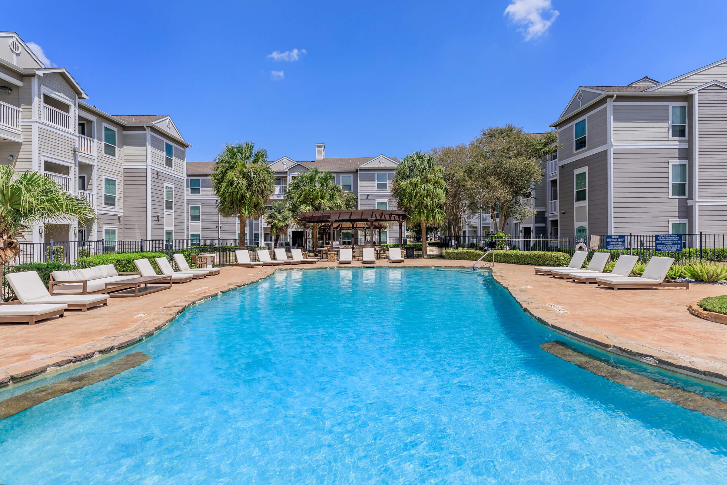A swimming pool surrounded by lounge chairs, with a shaded gazebo in the background. The pool is clear and inviting, set in a landscaped area with palm trees and modern apartment buildings under a blue sky.