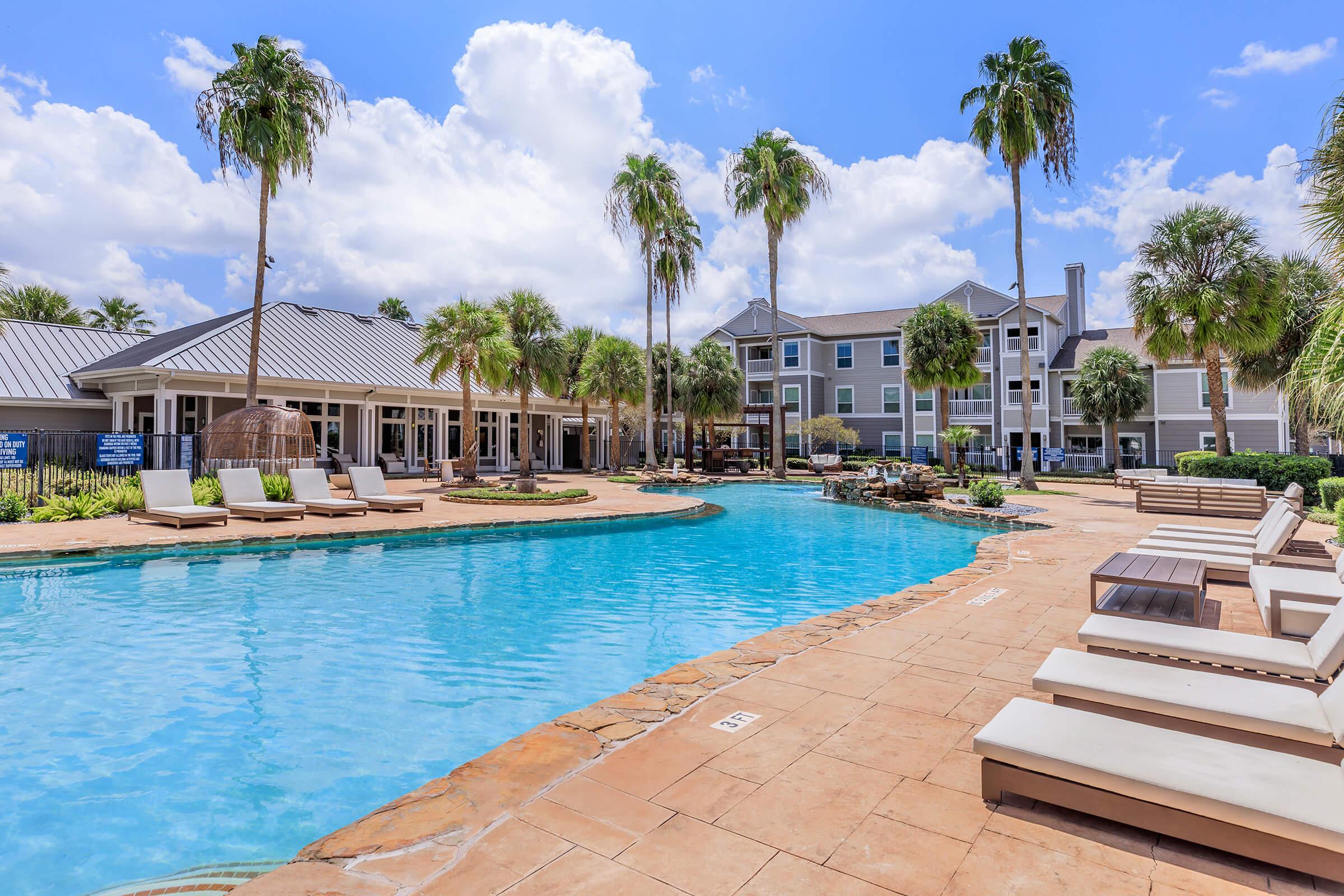 A beautiful resort-style pool surrounded by palm trees and lounge chairs. In the background, there are residential buildings under a bright blue sky with fluffy white clouds. The pool area features a stone waterfall, creating a serene and inviting atmosphere. Perfect for relaxation and leisure.