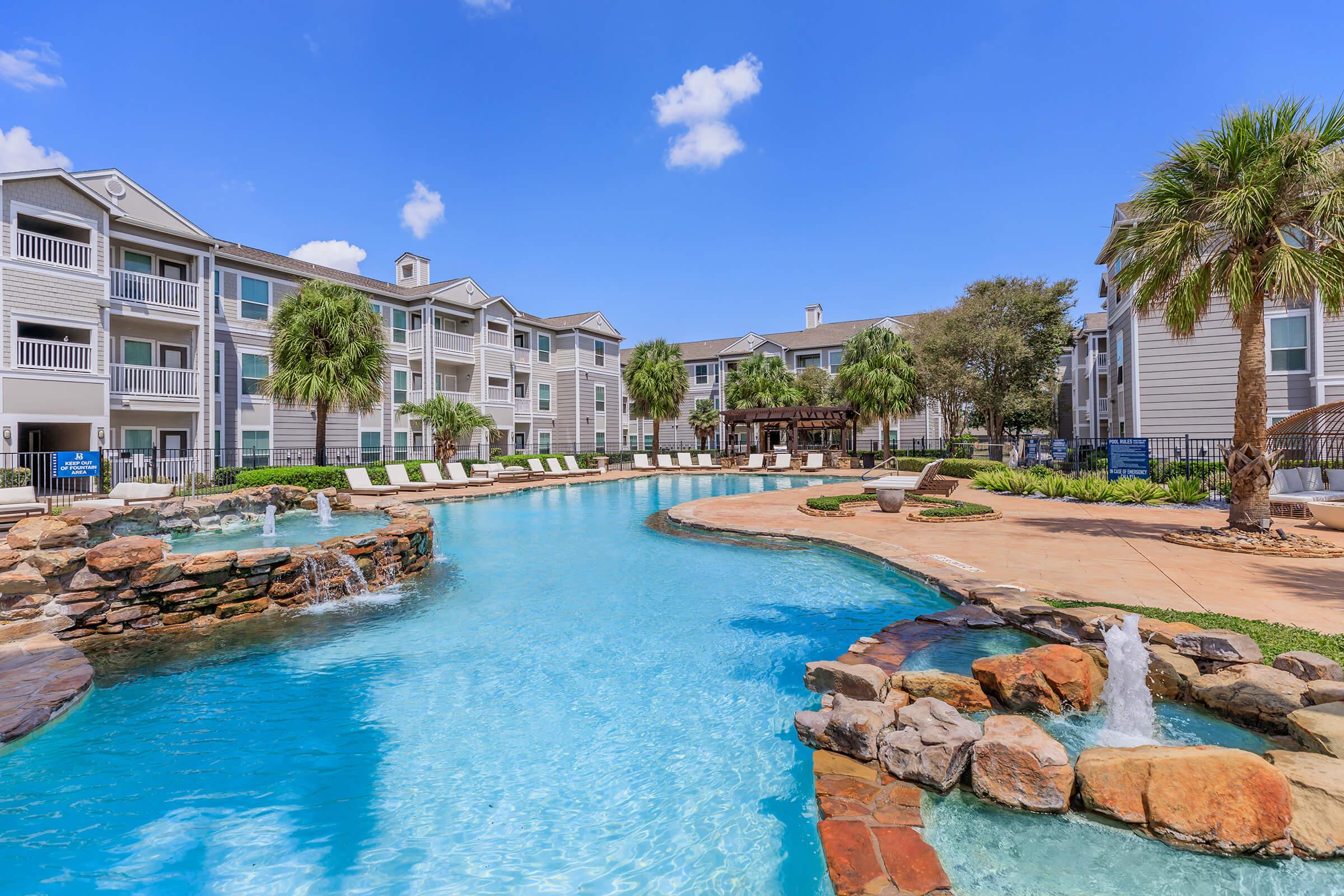 A serene outdoor swimming pool surrounded by rocky landscaping and palm trees, with lounge chairs positioned along the water. Modern apartment buildings are visible in the background under a clear blue sky. The scene conveys a relaxed, resort-like atmosphere.