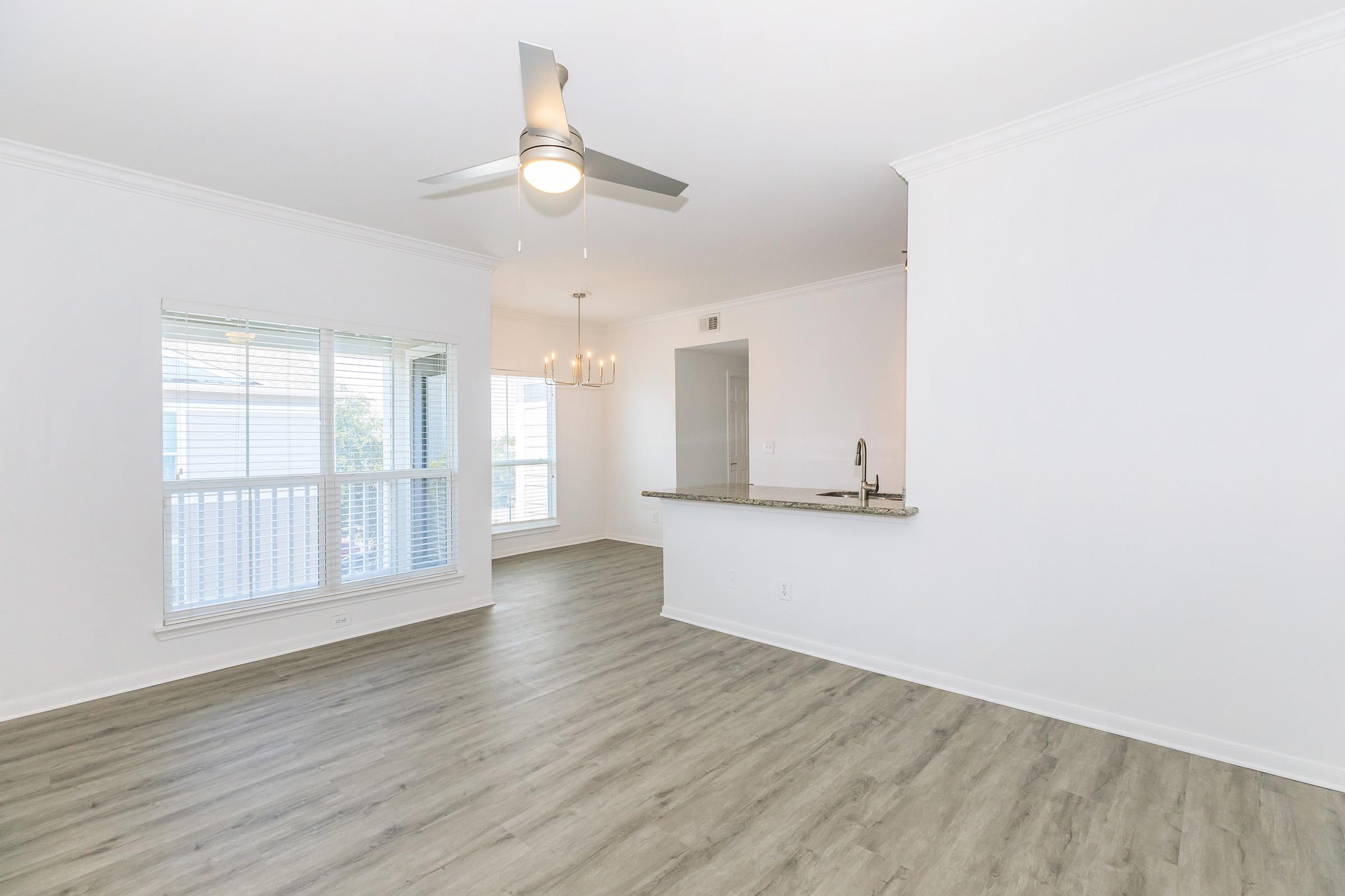 Bright, open living space featuring a light wood floor, large windows with white blinds, and a ceiling fan. A bar area with granite countertop separates the living room from the kitchen. The walls are painted white, providing a modern and airy feel to the room.