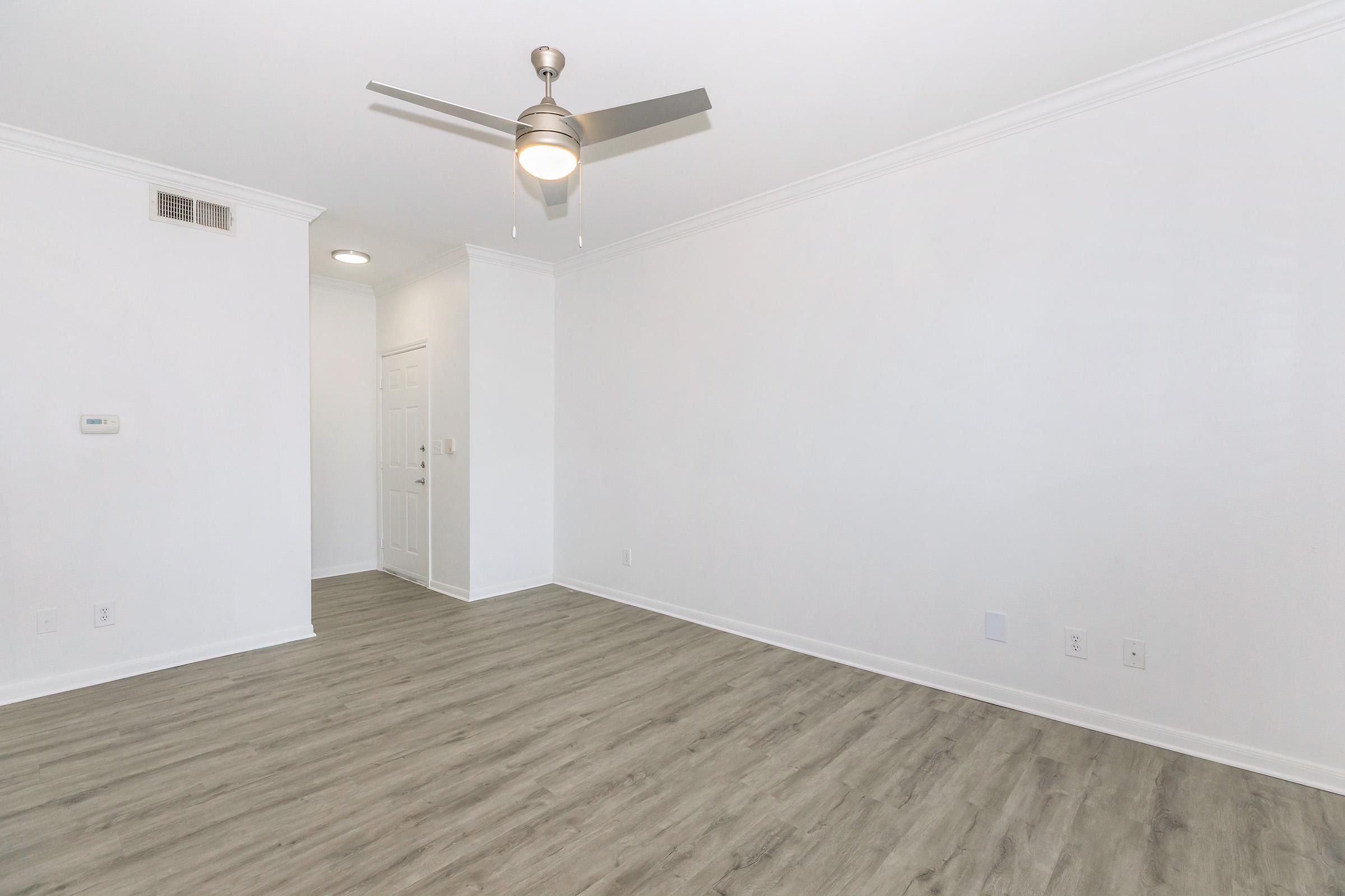 A spacious, empty room with light-colored walls and wood-like flooring. A ceiling fan is mounted on the ceiling, and there is a white door on one side leading to another room. The room is well-lit, creating a bright and airy atmosphere.