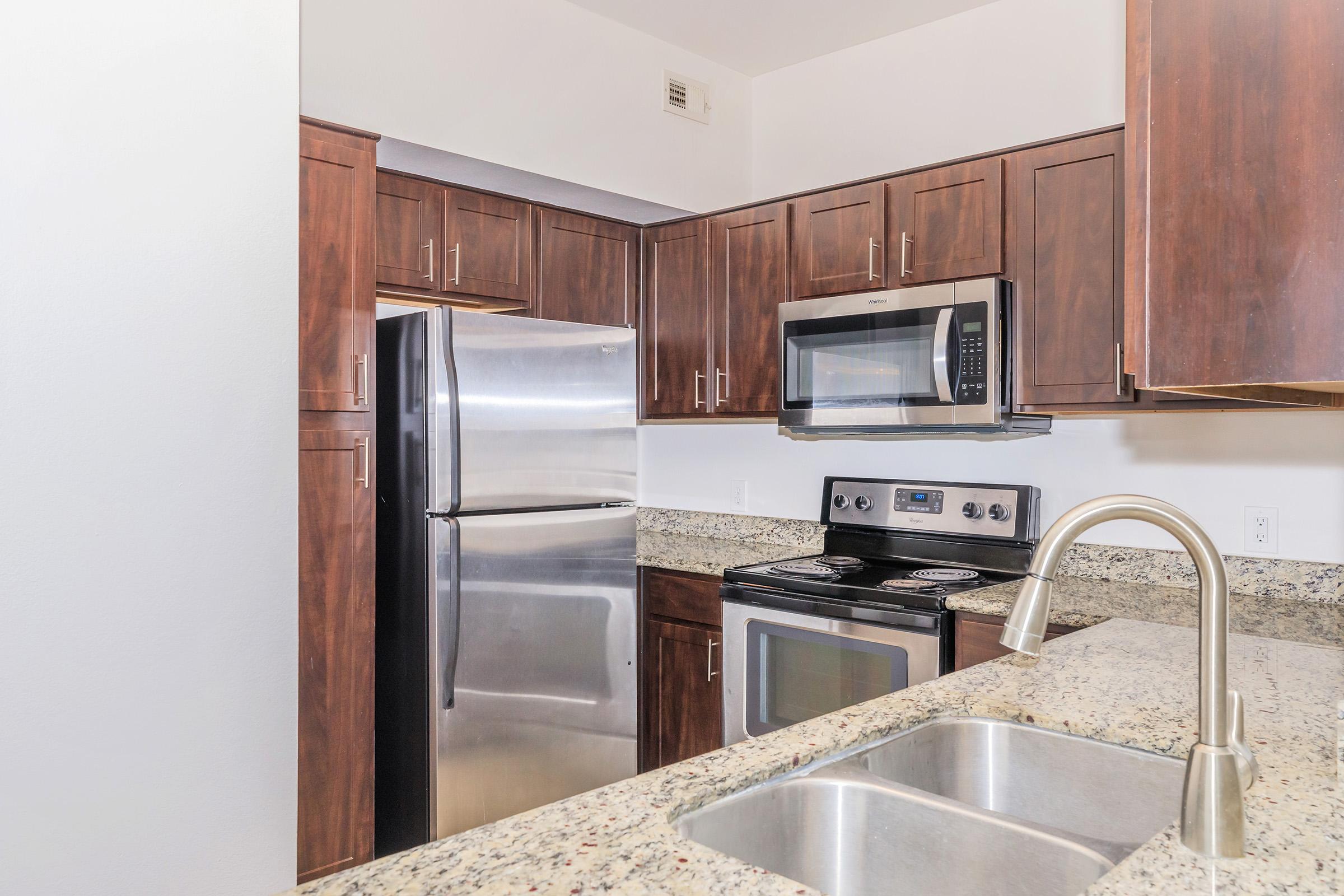 Modern kitchen featuring dark wooden cabinets, stainless steel appliances including a refrigerator and microwave, a smooth-top stove, and a dual-basin sink with a sleek faucet. The countertops are made of speckled granite, providing an elegant look in a well-lit space.