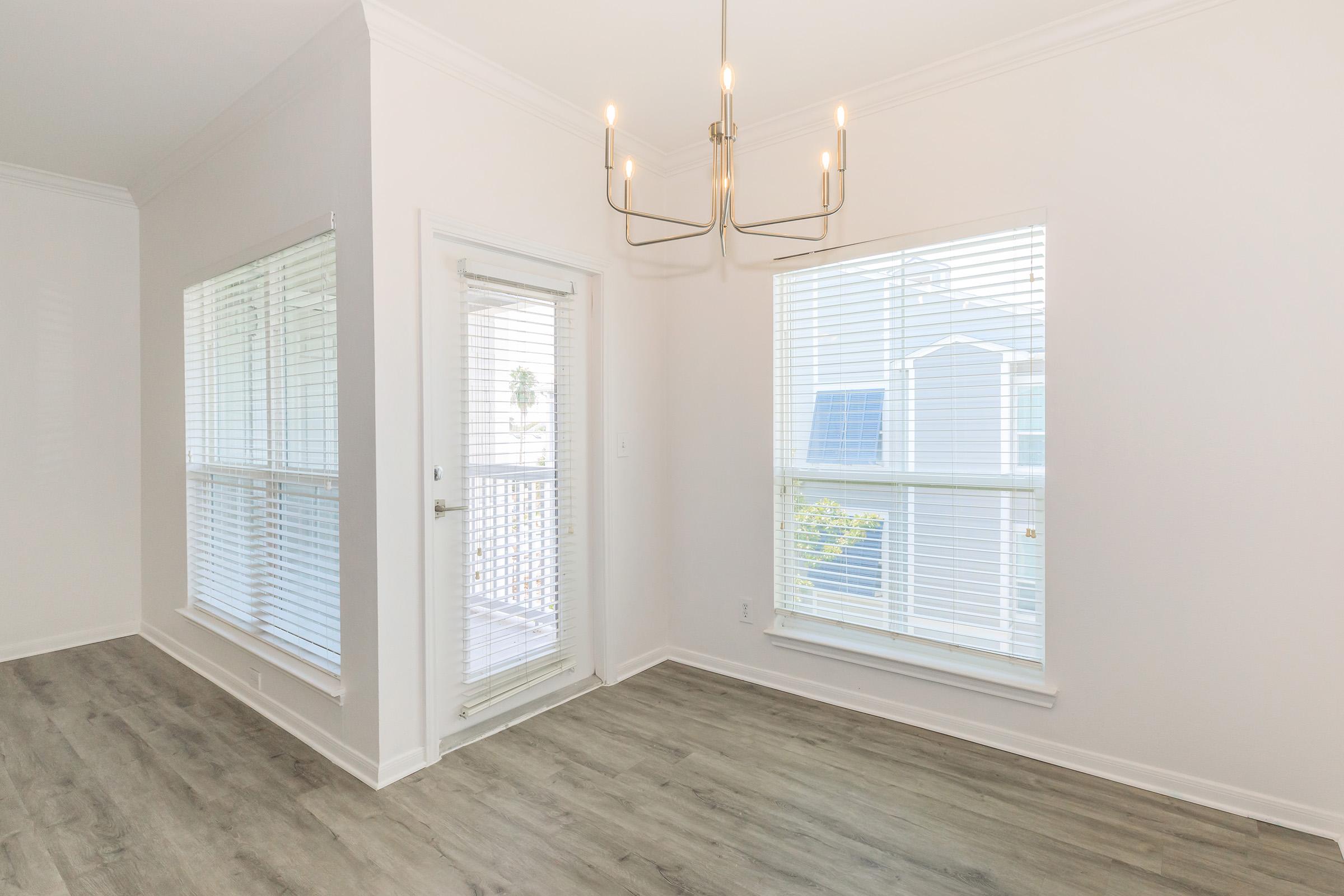 Bright, airy interior of a room featuring two large windows with white blinds. A sliding glass door leads to a balcony. The space has light-colored walls and a modern chandelier hanging from the ceiling. The flooring is light hardwood, creating a clean and inviting atmosphere.