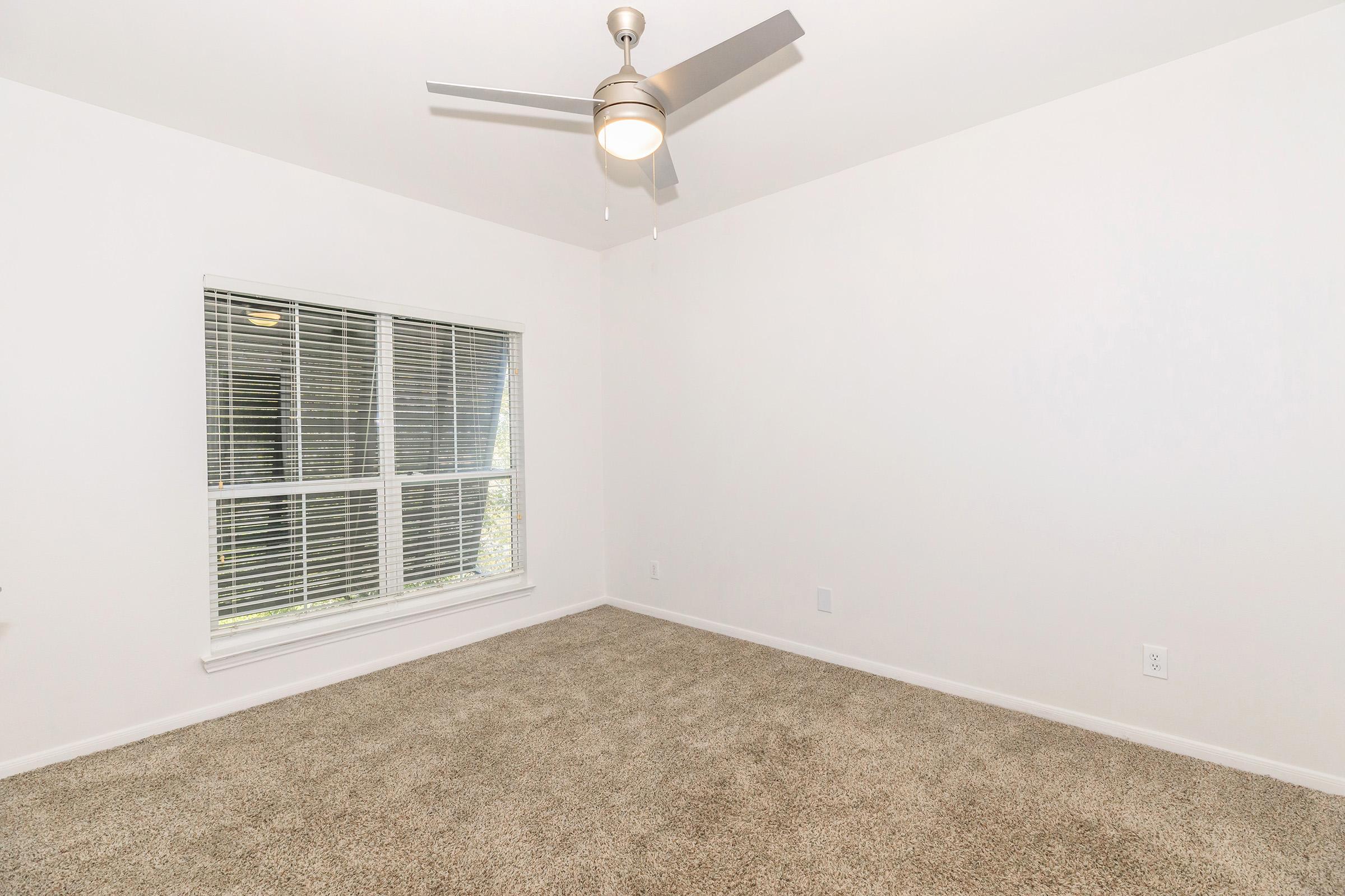 An empty room with light beige carpet, a ceiling fan, and a large window featuring closed blinds. The walls are painted white, creating a bright and airy atmosphere. The room is unfurnished, highlighting the spaciousness and potential for personal decoration.
