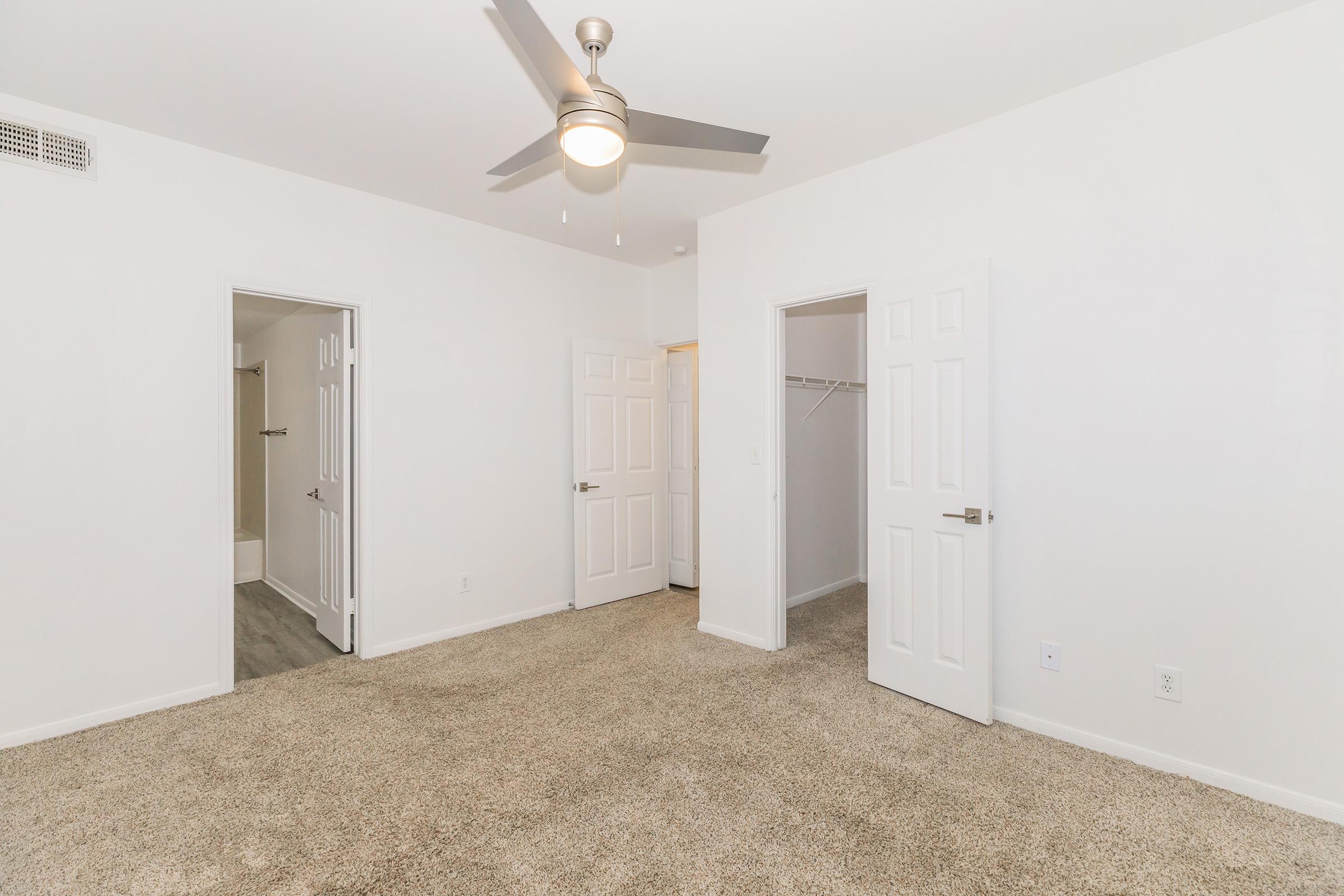 A well-lit, empty room featuring light-colored walls and a carpeted floor. There are two closed doors on the left leading to separate spaces, while the door on the right is slightly ajar, revealing a closet. A modern ceiling fan is installed, enhancing the room's spacious feel.