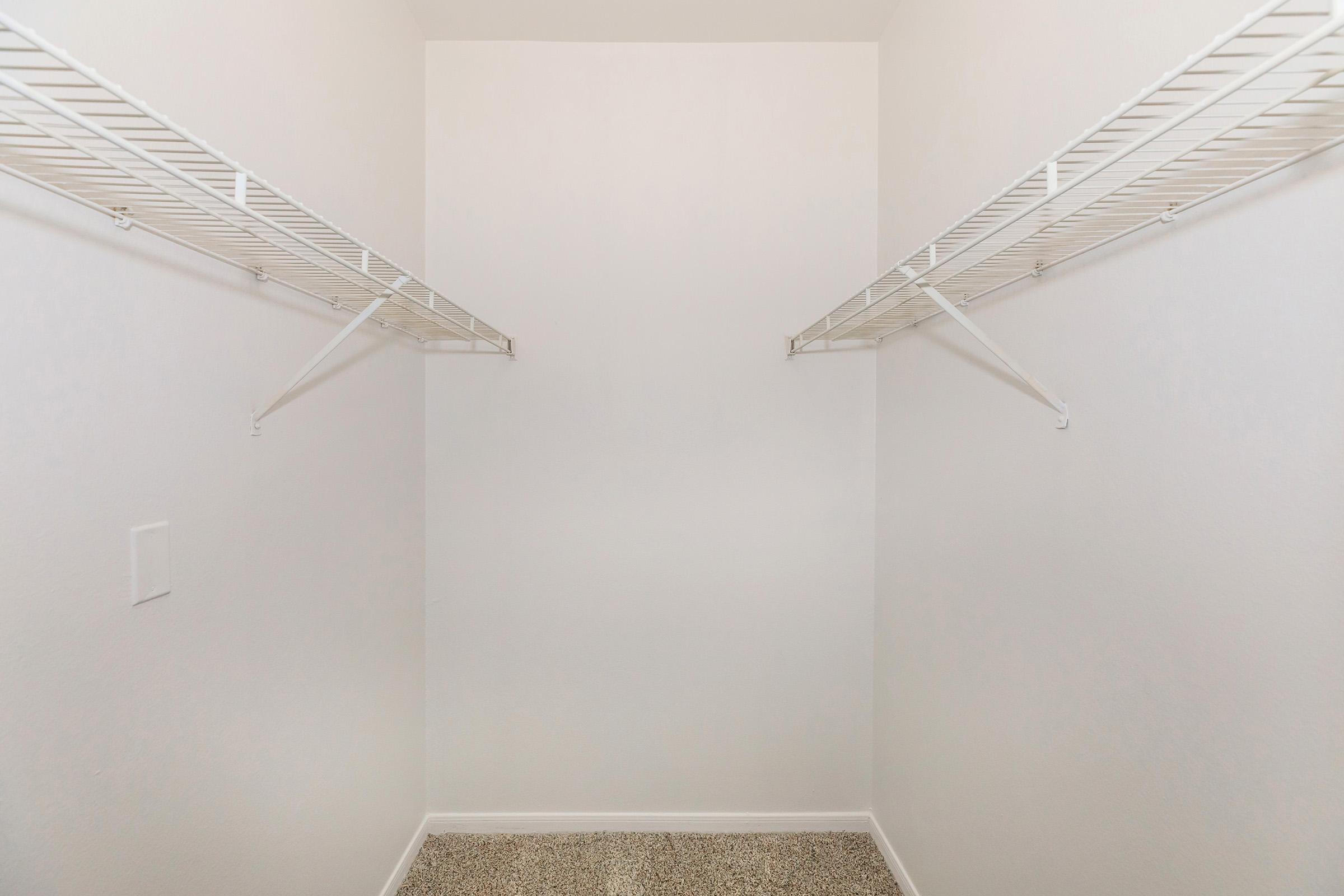 Empty walk-in closet featuring white wire shelving on both sides against plain white walls, with a beige carpeted floor. The space is uncluttered and minimalistic, lacking any stored items or decor.