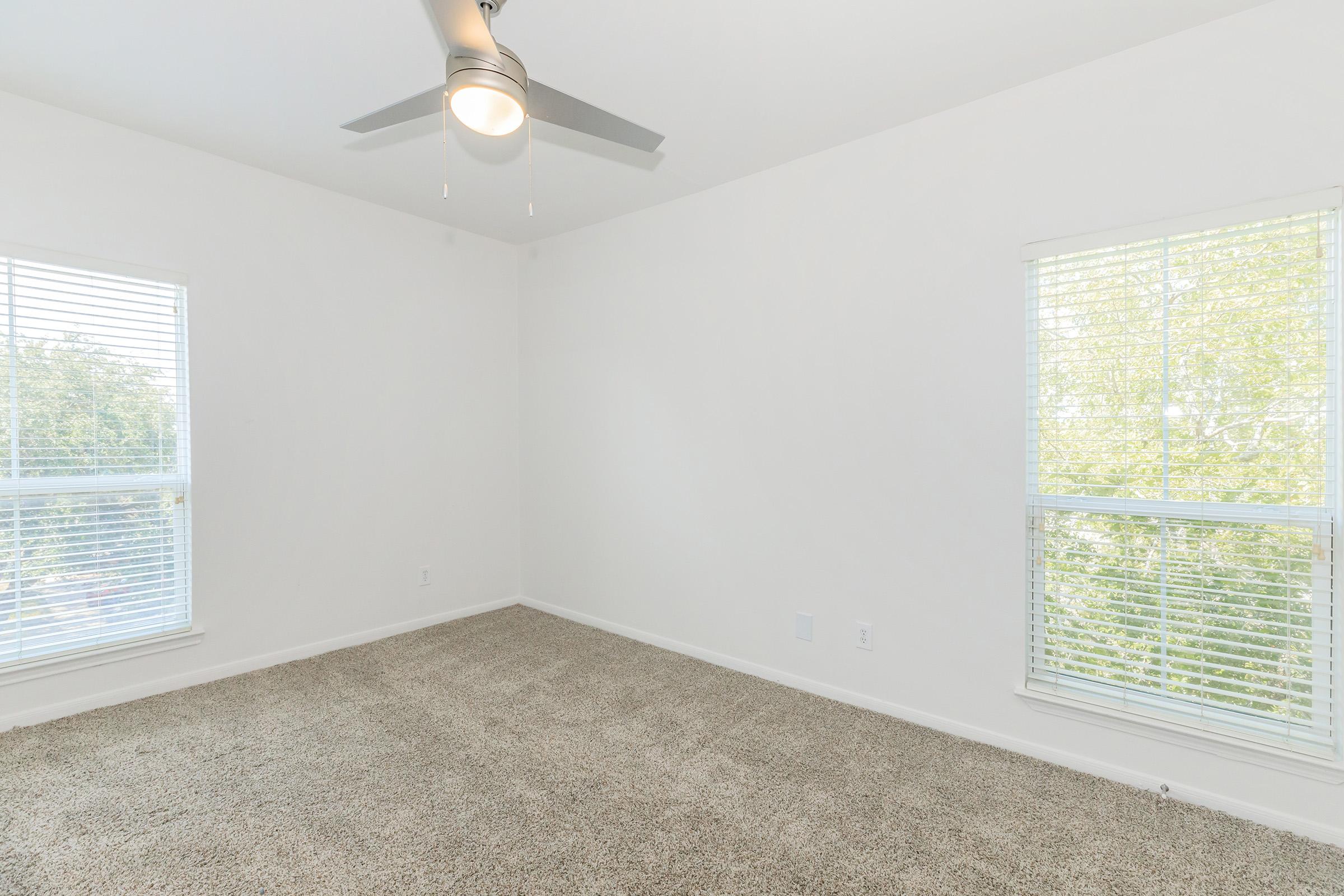 Empty room with light beige carpet, white walls, and a ceiling fan. There are two windows with white blinds letting in natural light, showing greenery outside. The space appears clean and well-lit, suitable for various uses such as a bedroom or office.