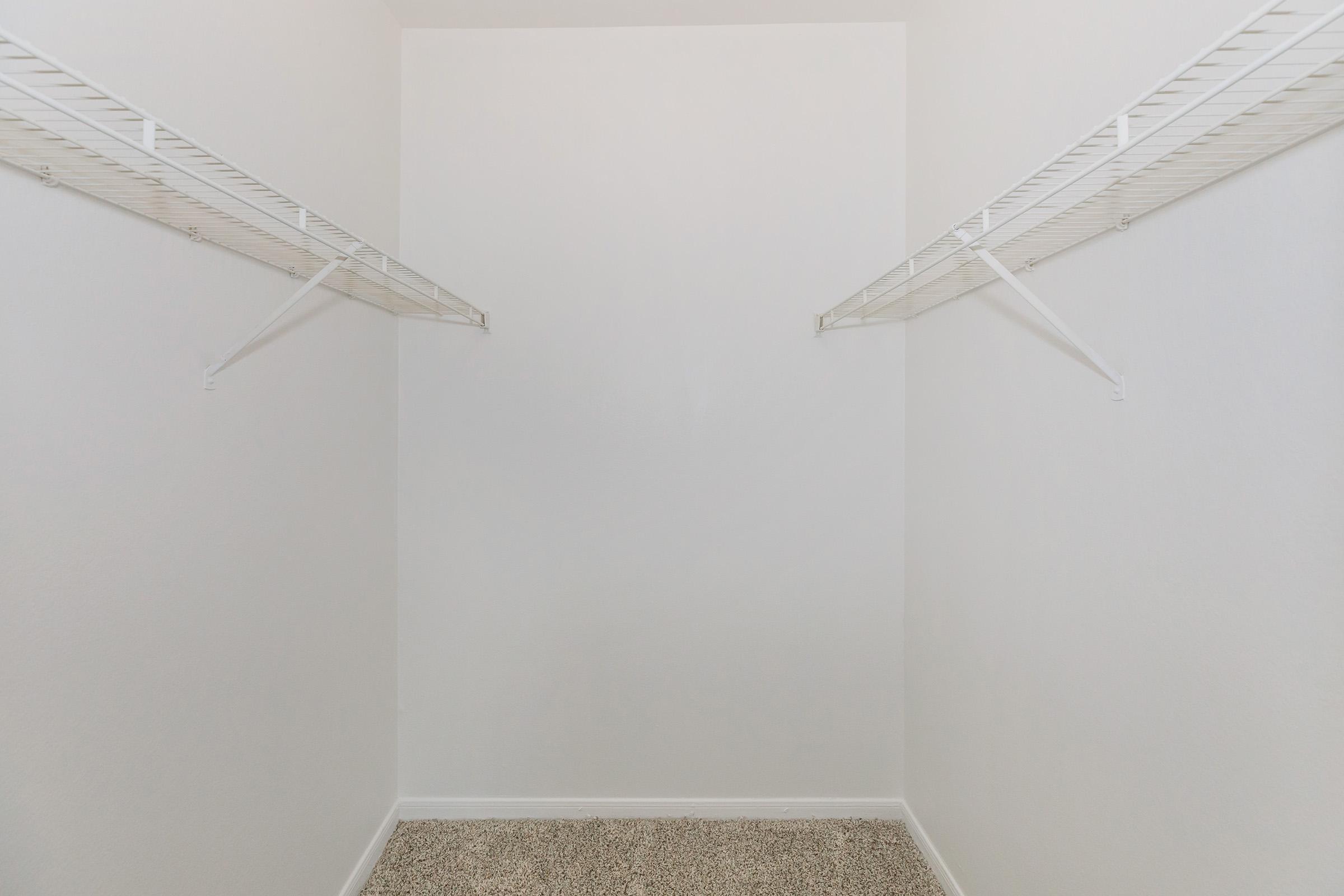 Empty closet with light-colored walls and a carpeted floor. Two wire shelves are mounted on the walls, one on each side, providing storage space. The overall space is minimal and devoid of any items or decorations, emphasizing a clean and simple design.
