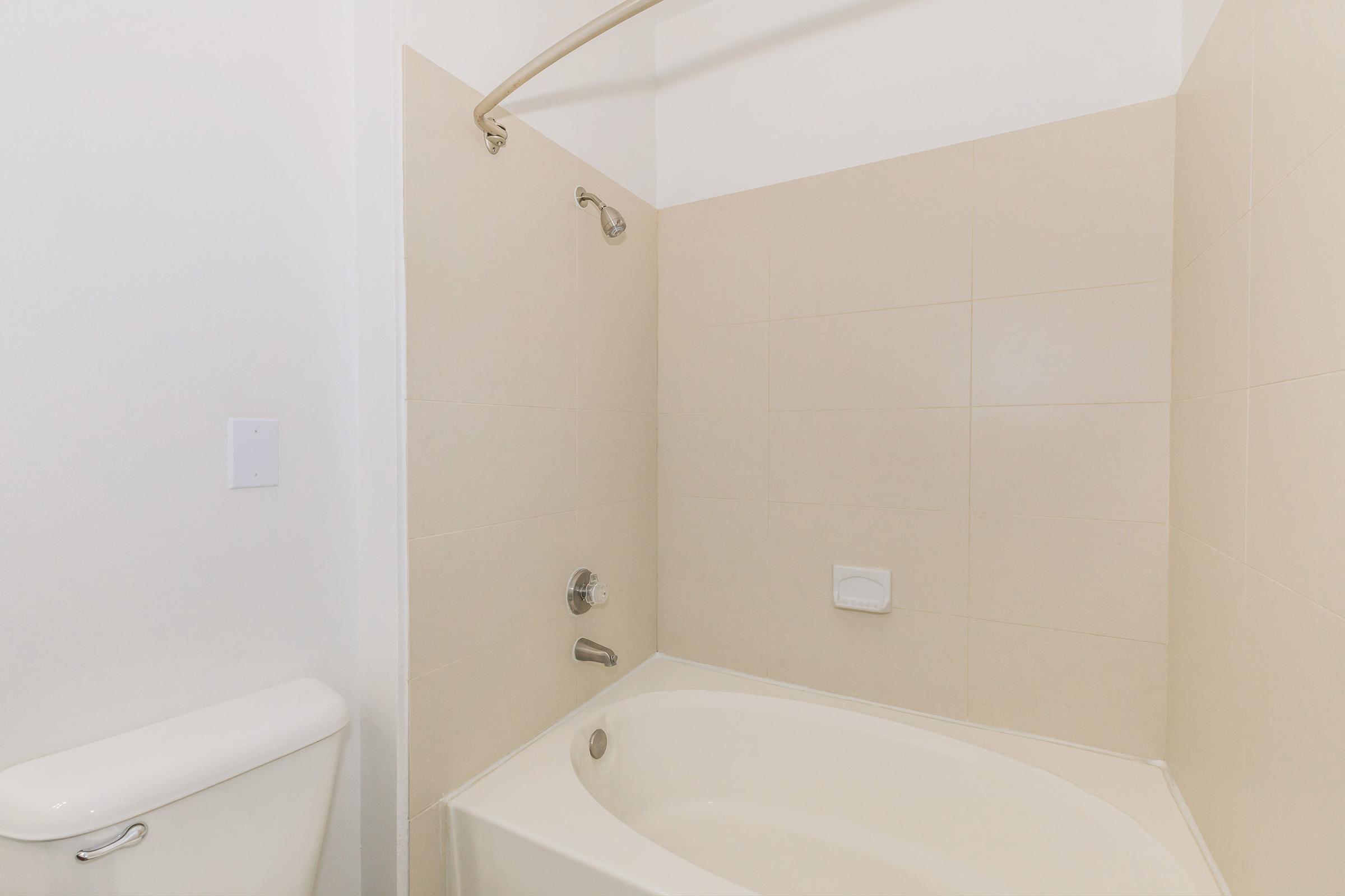 A clean bathroom featuring a bathtub with a curved shower rod, a wall-mounted faucet, and beige tiled walls. There is a white toilet adjacent to the tub, and the overall design is simple and modern with a neutral color palette.