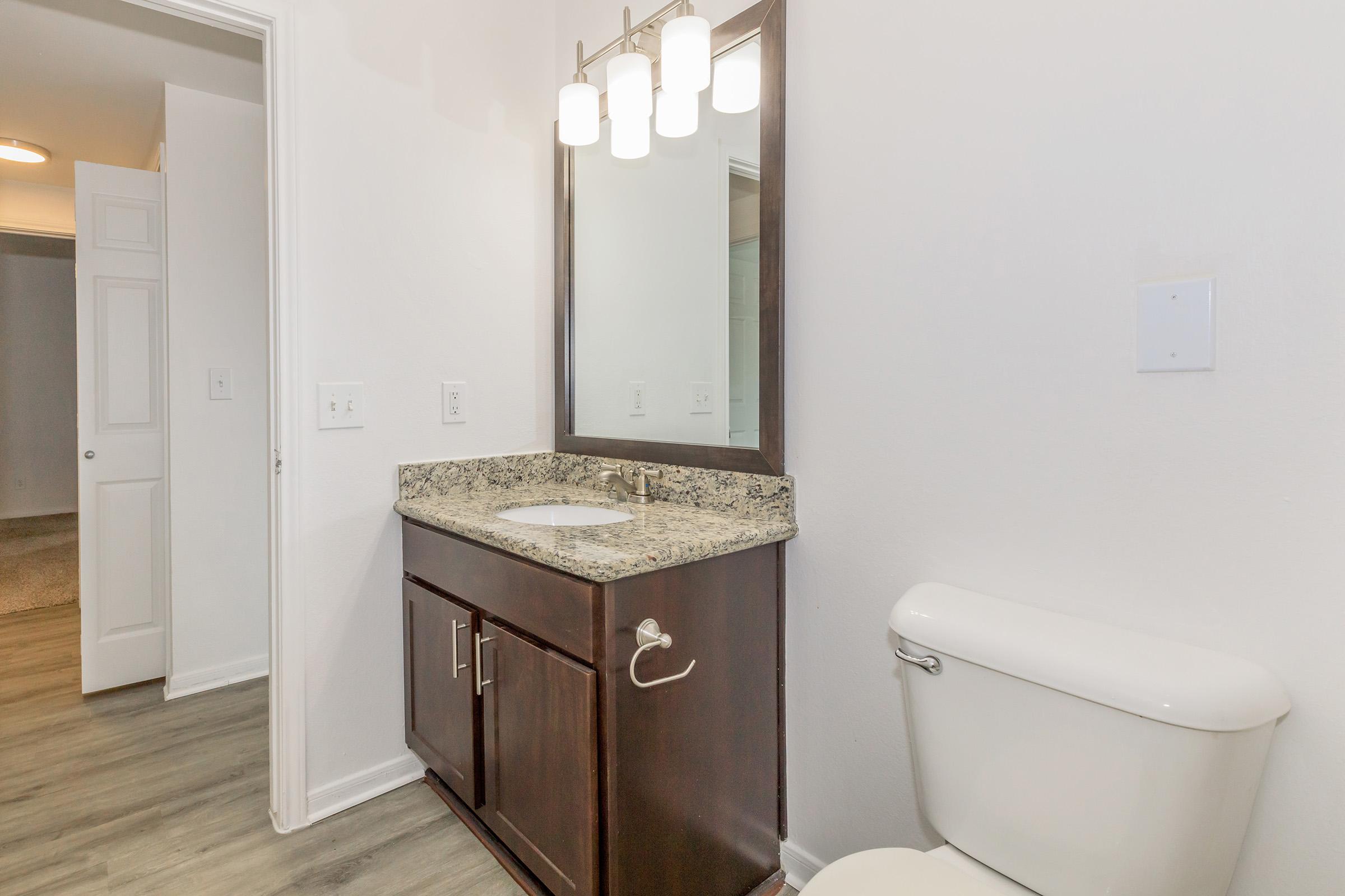 A modern bathroom featuring a dark wood vanity with a granite countertop, a rectangular mirror above it, and three hanging light fixtures. There is a white toilet beside the vanity. The walls are painted white, and the flooring is a light wood texture. A doorway is visible in the background leading to another room.