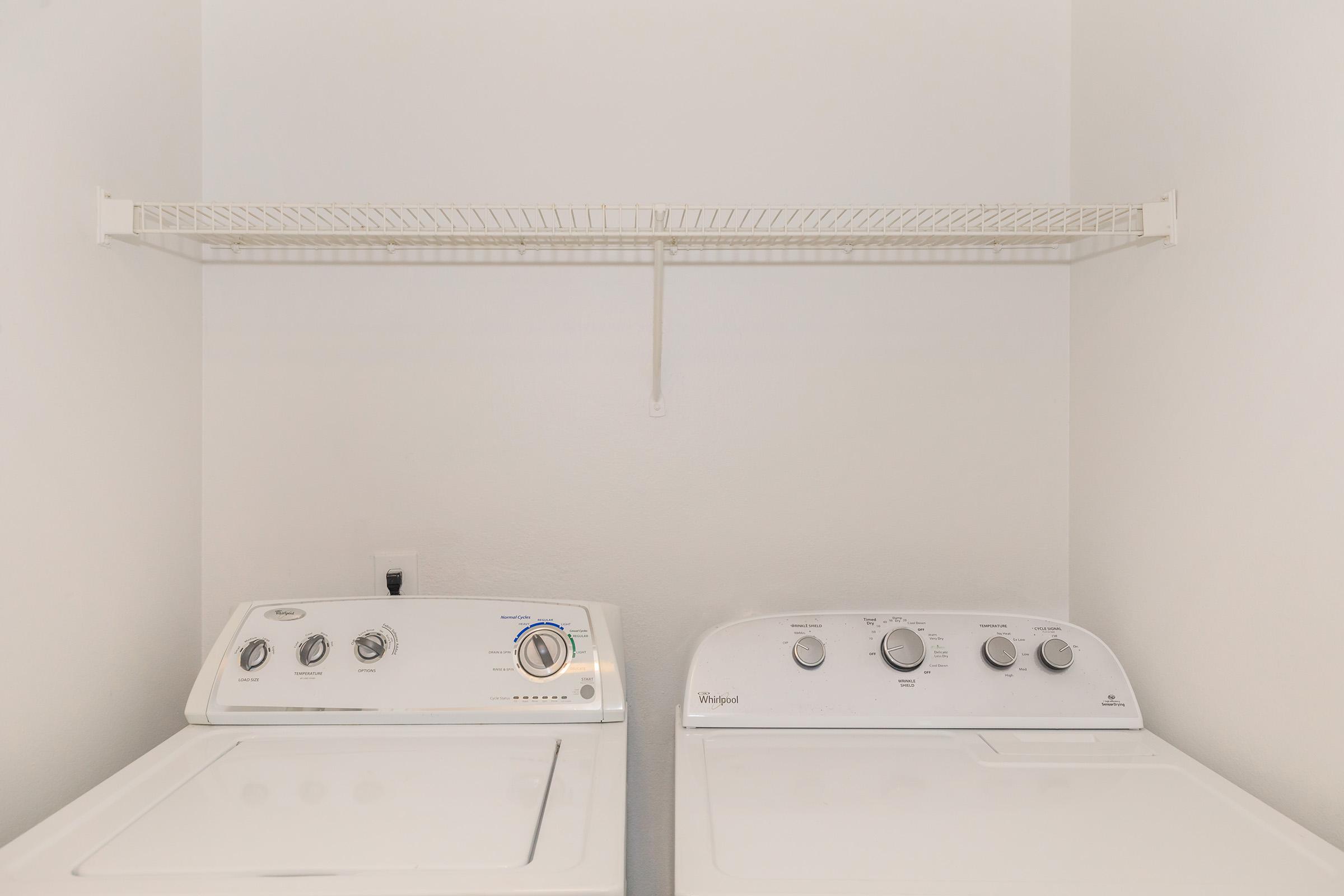 A clean laundry room featuring a washing machine and a dryer side by side. Above them, there is a white wire shelf mounted on the wall for storage. The walls are painted white, and the floor is visible, creating a bright and organized space.