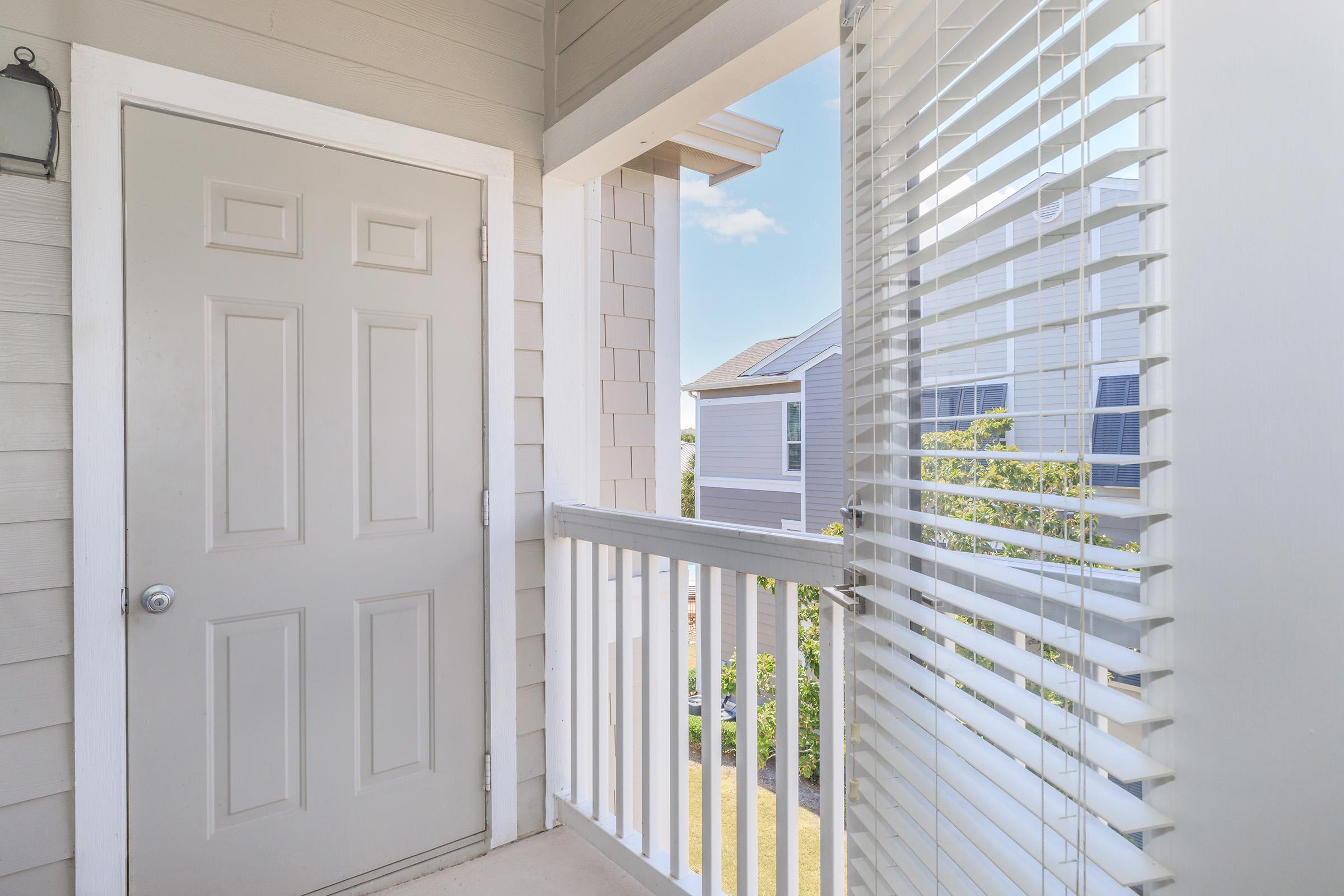 An exterior view from a balcony showing a closed gray door, white railings, and partially opened white blinds. In the background, two houses are visible against a clear blue sky. The scene captures a bright and inviting outdoor space.