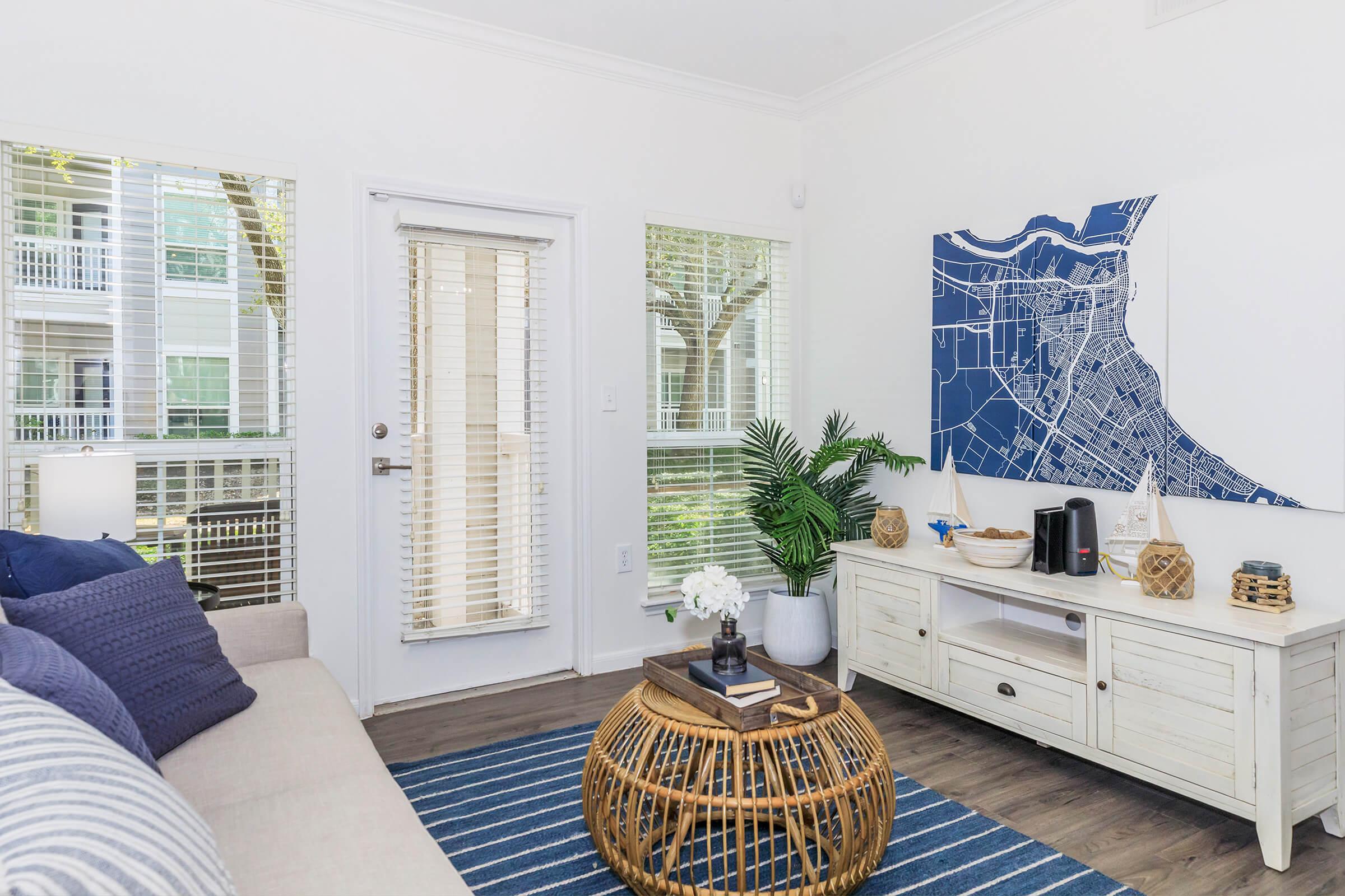 A cozy living room featuring a light-colored sofa with blue throw pillows, a round woven coffee table, and a stylish white media console. The room has large windows with blinds, providing natural light, and a map artwork on the wall. Indoor plants add a touch of greenery to the space.