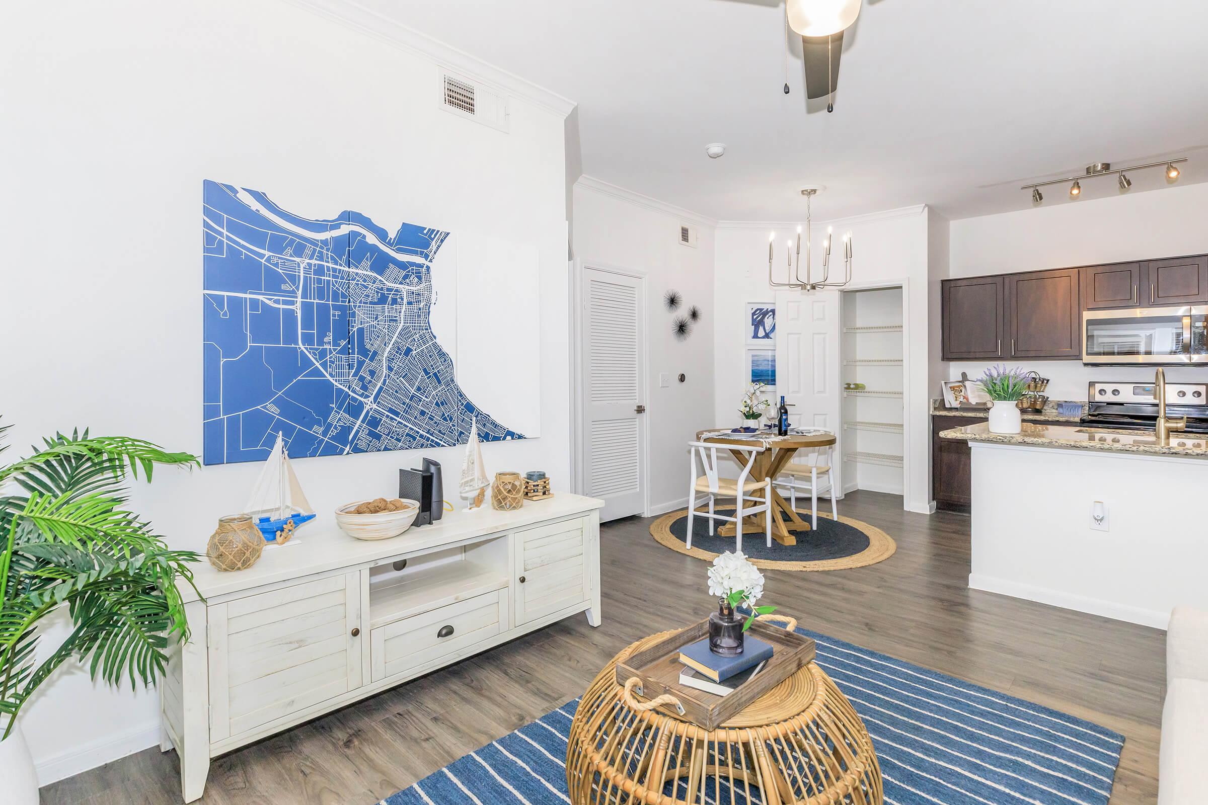 A modern living room featuring a large blue map artwork on the wall, a light-colored wooden media console with decorative items, a round woven coffee table, a striped blue and white area rug, and a kitchen area in the background with dark wood cabinetry and a dining table set for four.