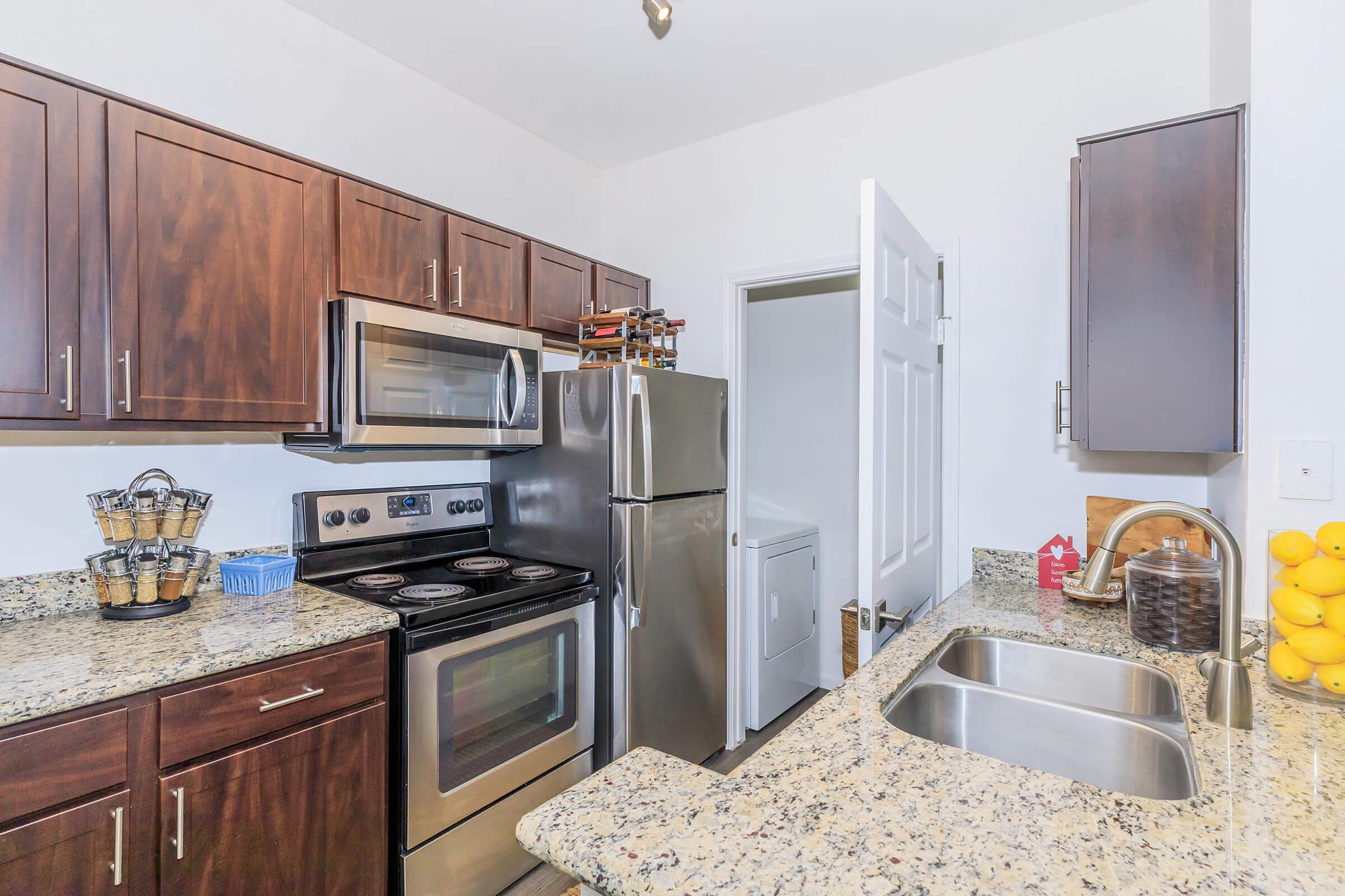 A modern kitchen featuring wooden cabinets, stainless steel appliances including a stove, refrigerator, and microwave. The countertops are made of granite, and there's a double sink. In the background, a laundry area is visible through an open door, along with a basket holding lemons.