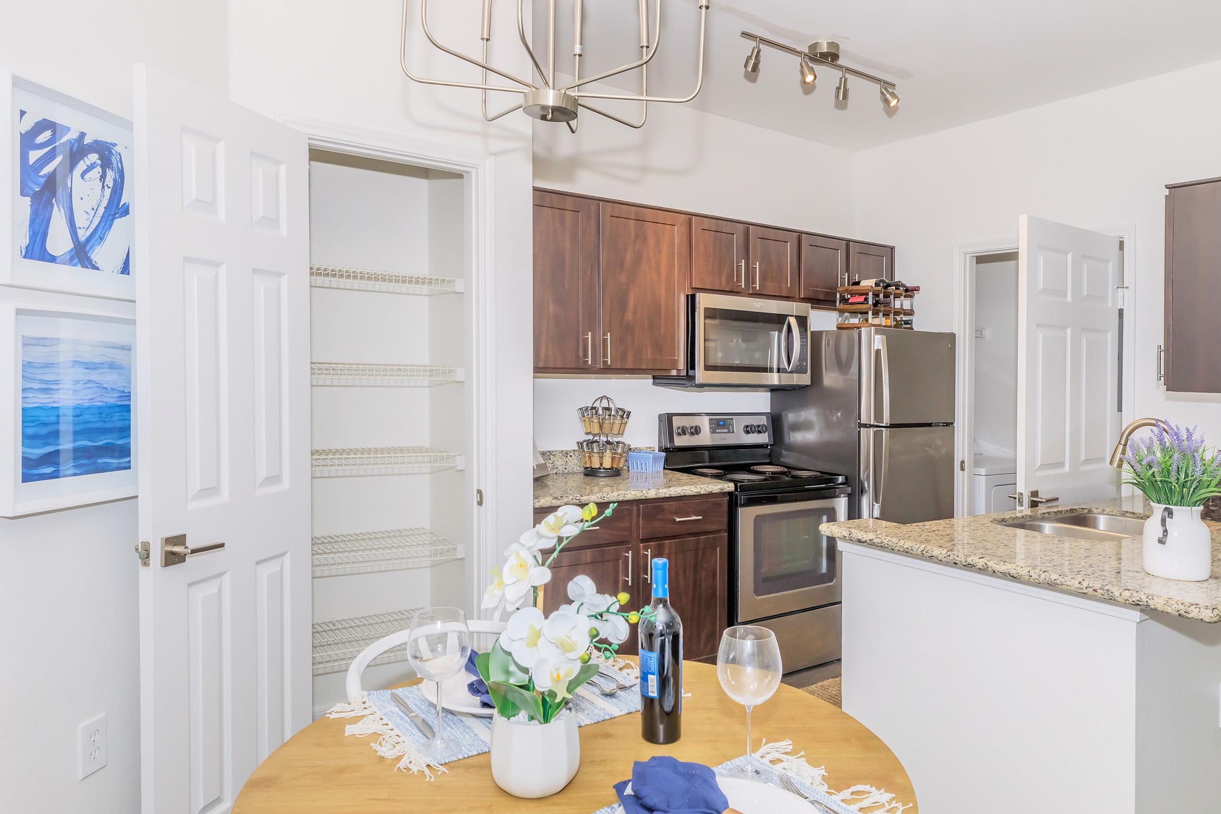 A modern kitchen featuring dark wood cabinets, stainless steel appliances, and granite countertops. There is a dining table adorned with a vase of flowers and wine glasses. Pantry doors are open, showcasing additional storage. Bright lighting adds warmth to the space.