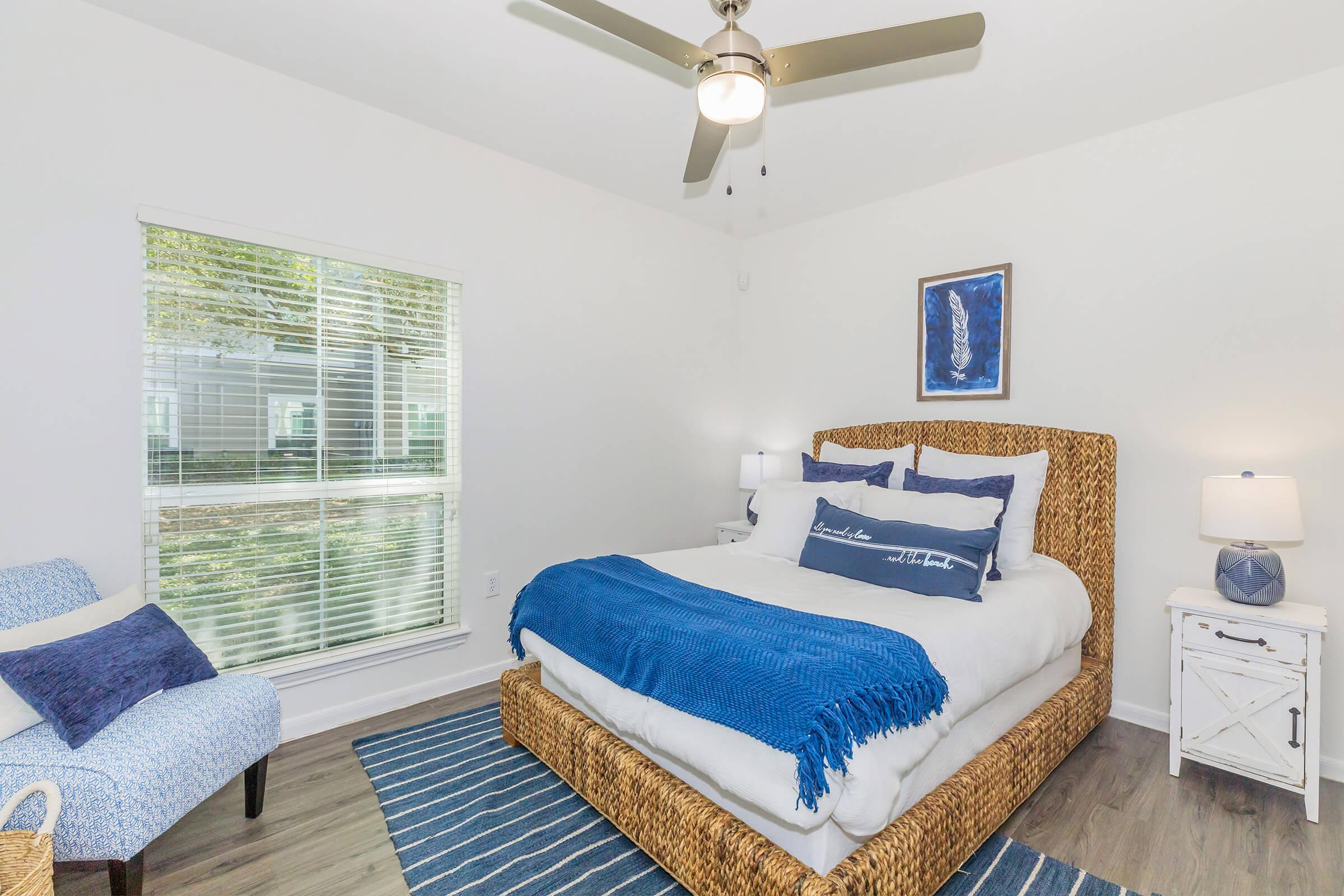 A bright bedroom featuring a large bed with a woven headboard and blue bedding. There is a blue throw blanket on the bed and a striped area rug beneath. A small armchair is in the corner next to a window with blinds, allowing natural light in. The room has light walls and a ceiling fan.