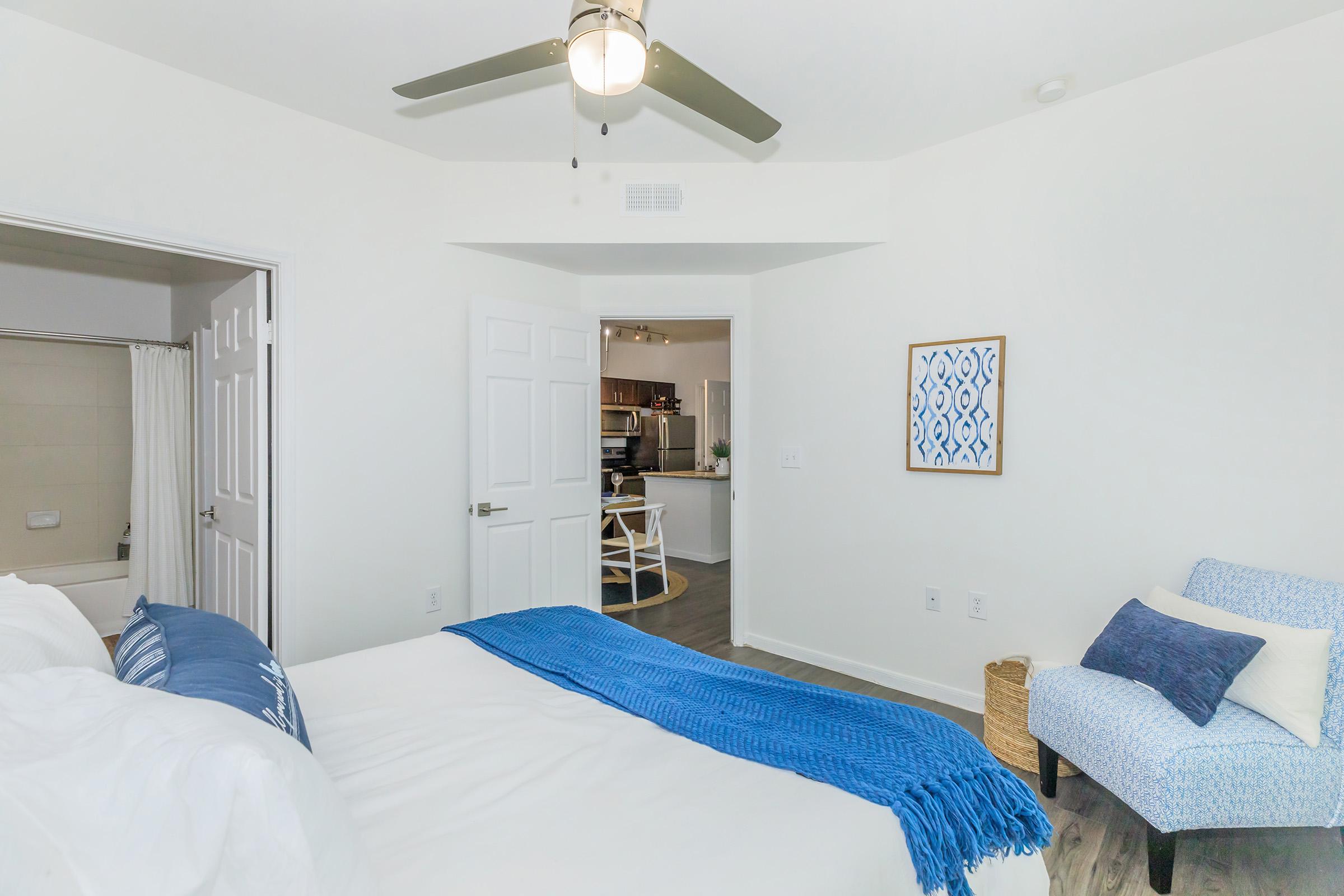 A bright and modern bedroom featuring a white bed with a blue throw blanket, a ceiling fan, and a small seating area with a light blue chair. The room has white walls, a doorway leading to a kitchen, and a framed blue wall art piece. Natural light illuminates the space, creating a welcoming atmosphere.