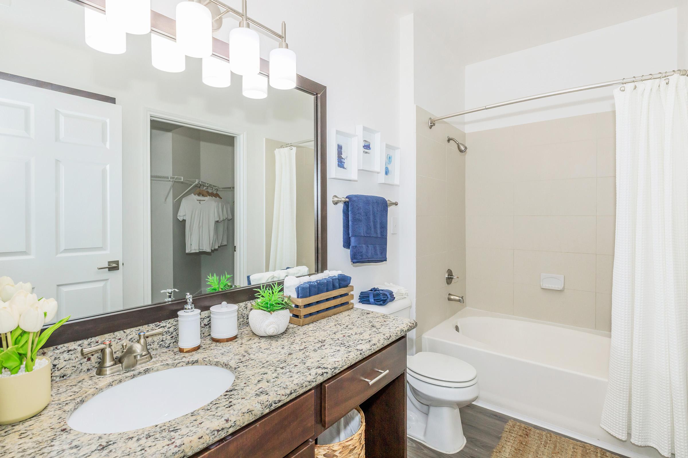 A well-lit bathroom featuring a granite countertop with a sink, decorative plants, and neatly arranged toiletries. The space includes a shower-tub combination with a white curtain, a towel rack with blue towels, and a glimpse of a closet in the background with white clothing.