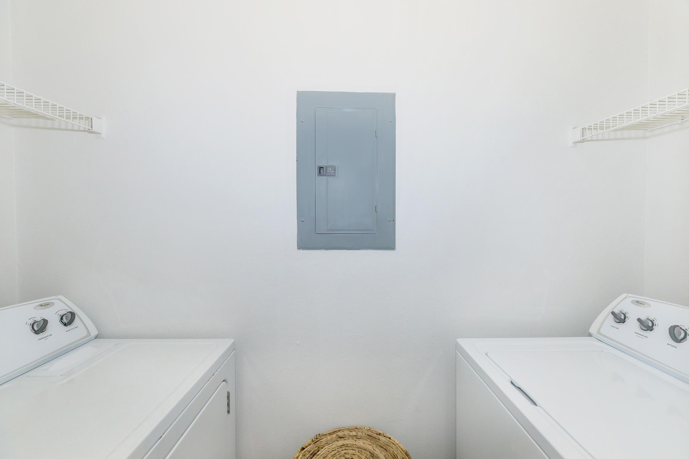 A clean, well-lit laundry room featuring two white washing machines. The machines are positioned side by side against a plain white wall. Above them, there are wire shelves for storage. A gray electrical panel is mounted on the wall in the center, adding utility to the space.