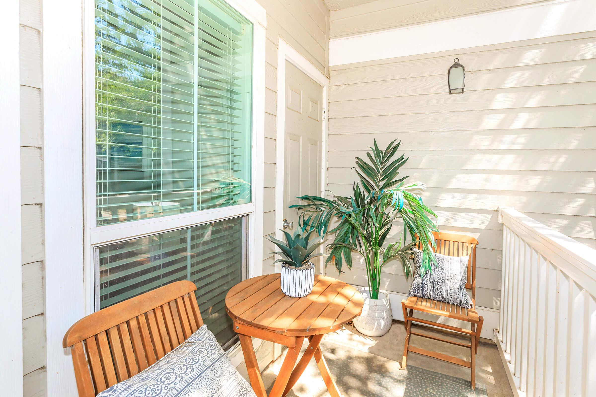 A cozy balcony featuring two wooden chairs and a small round table. A potted plant adds a touch of greenery, while sunlight filters through the window's blinds, creating a warm and inviting atmosphere. The walls are pale, enhancing the bright and airy feel of the space.