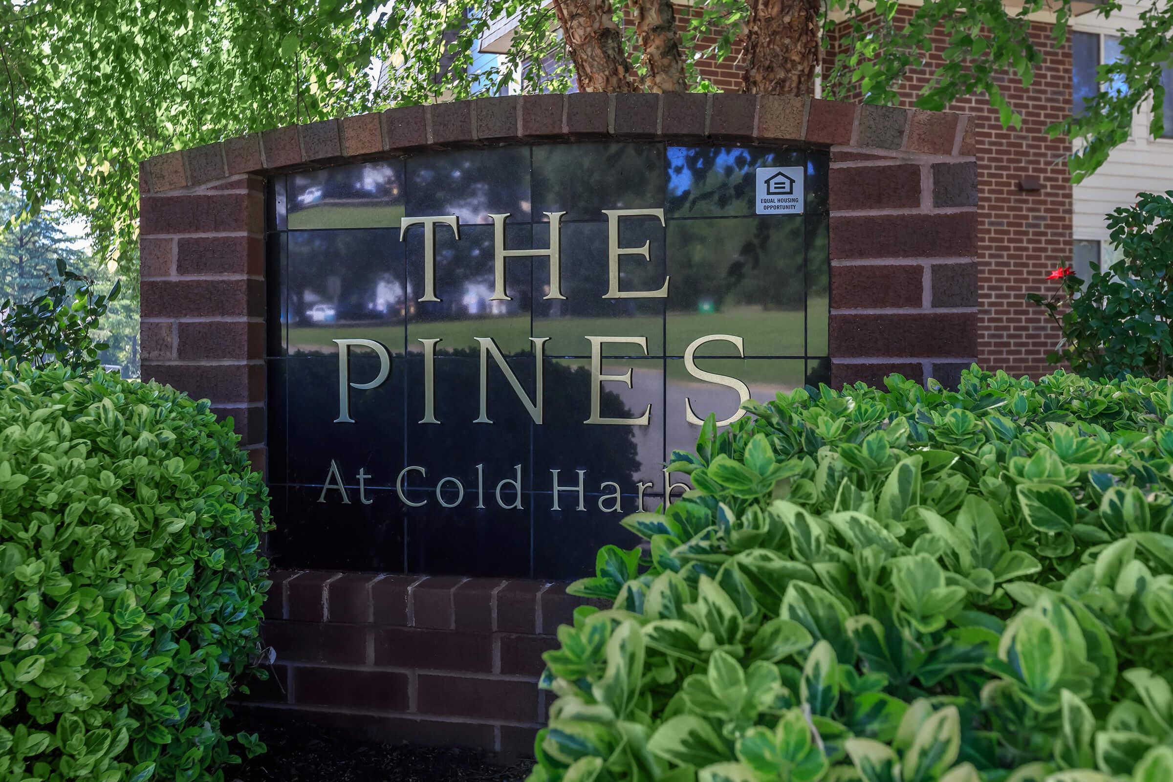 Sign that reads "The Pines At Cold Harbor," surrounded by green shrubs and trees, located near a building. The sign is made of dark stone with a modern design, contrasting with the vibrant greenery.