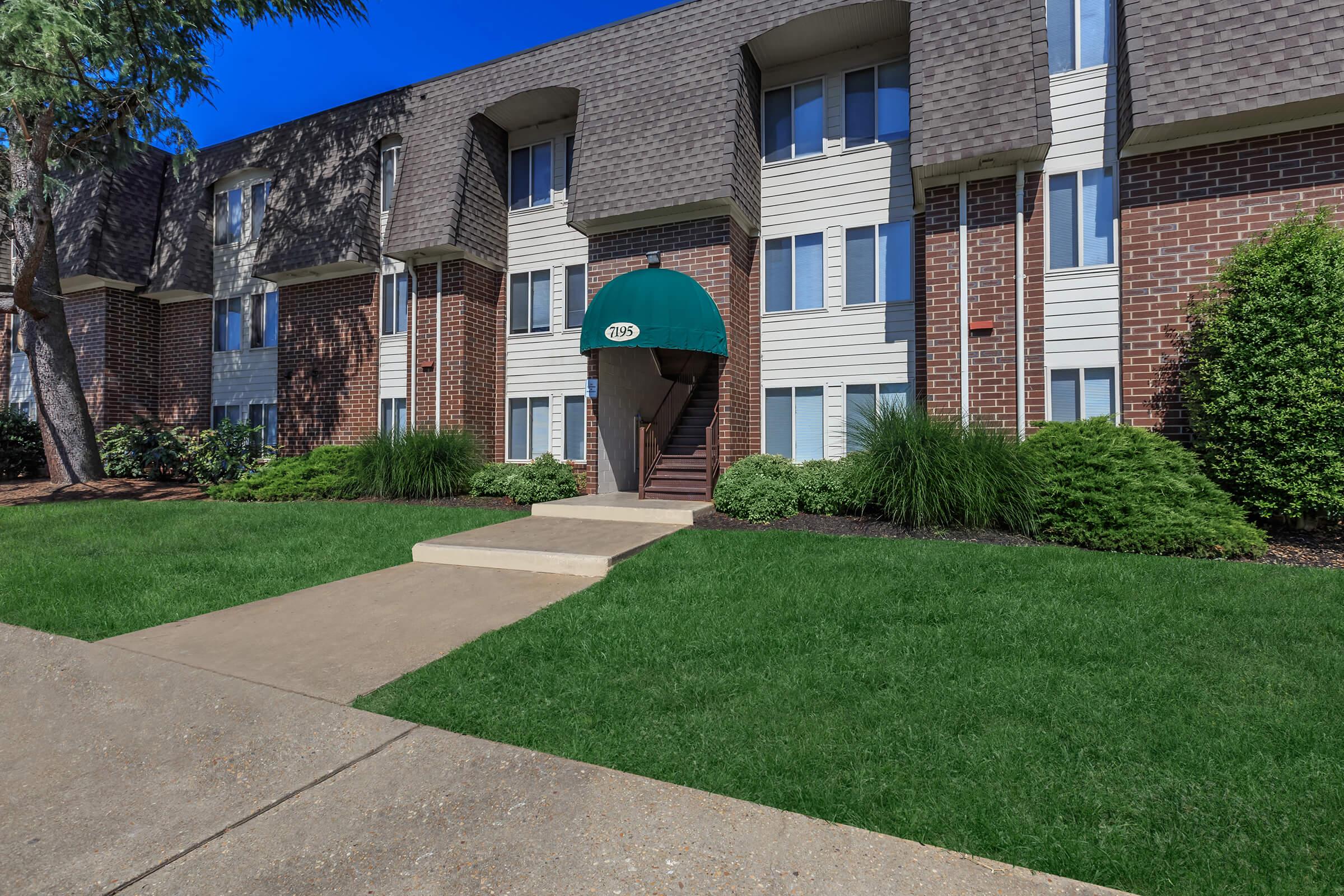A well-maintained apartment complex featuring brick and siding exterior. The entrance has a green awning and stairs leading up to the door. Surrounding the building are neatly trimmed shrubs and a manicured lawn, with a clean concrete pathway leading to the entrance. Bright blue sky in the background.