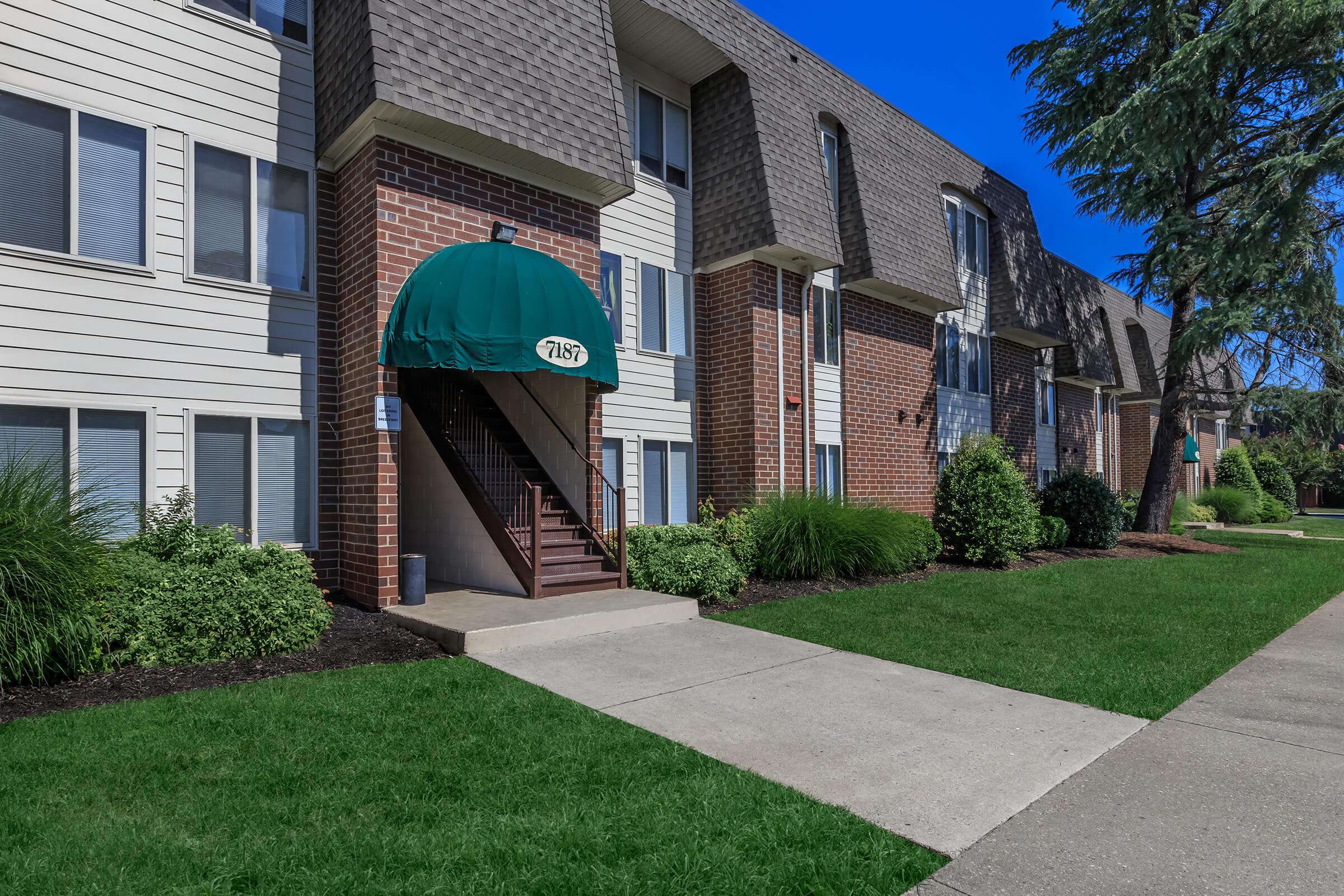 Exterior view of a multi-story apartment building featuring a green awning above the entrance. There are landscaped lawns with shrubs, a concrete walkway leading to the entrance, and clear blue sky in the background.