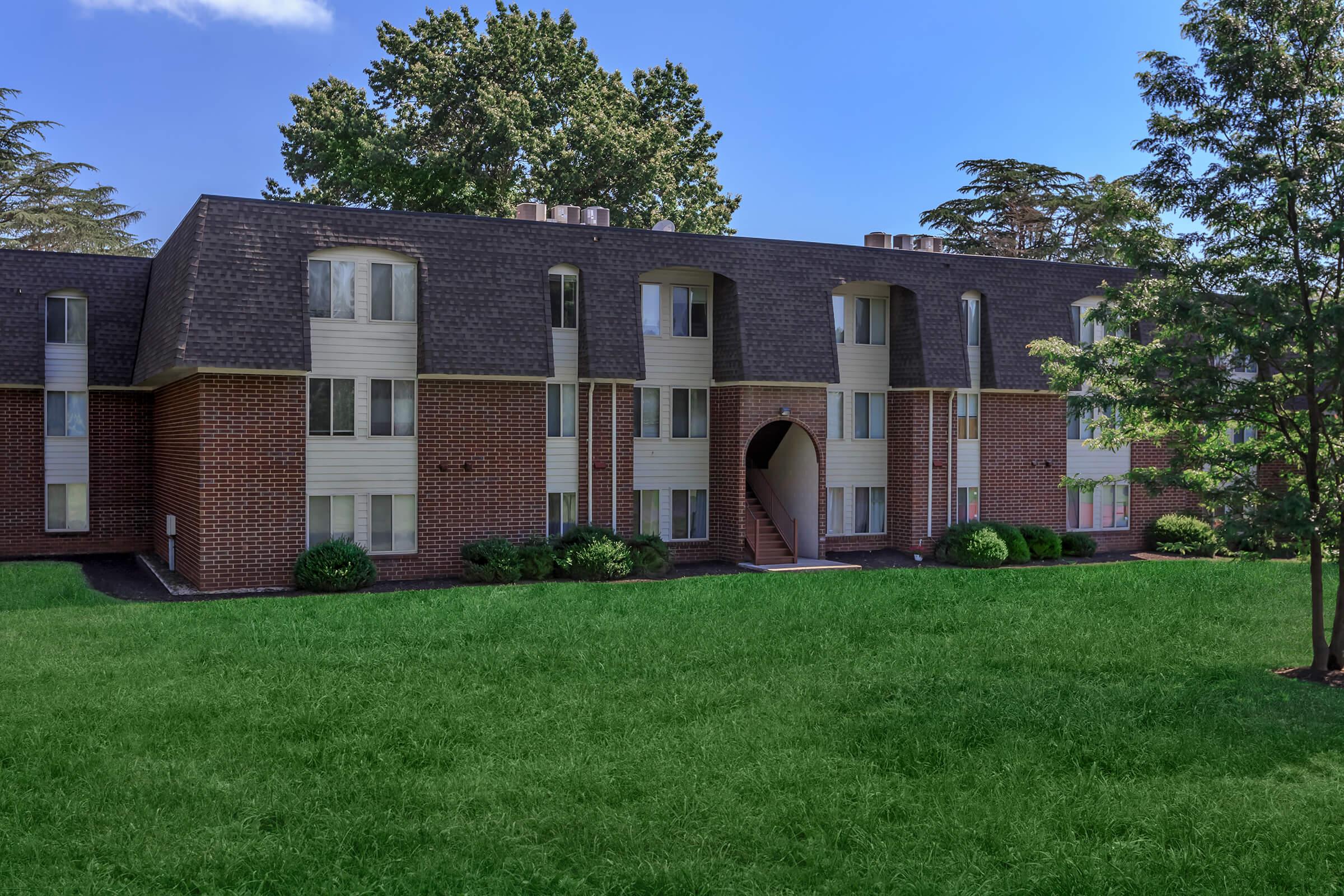 A multi-story brick apartment building with multiple windows and a central entrance, surrounded by green grass and a few shrubs. The sky is clear with a few trees in the background, creating a serene suburban atmosphere.