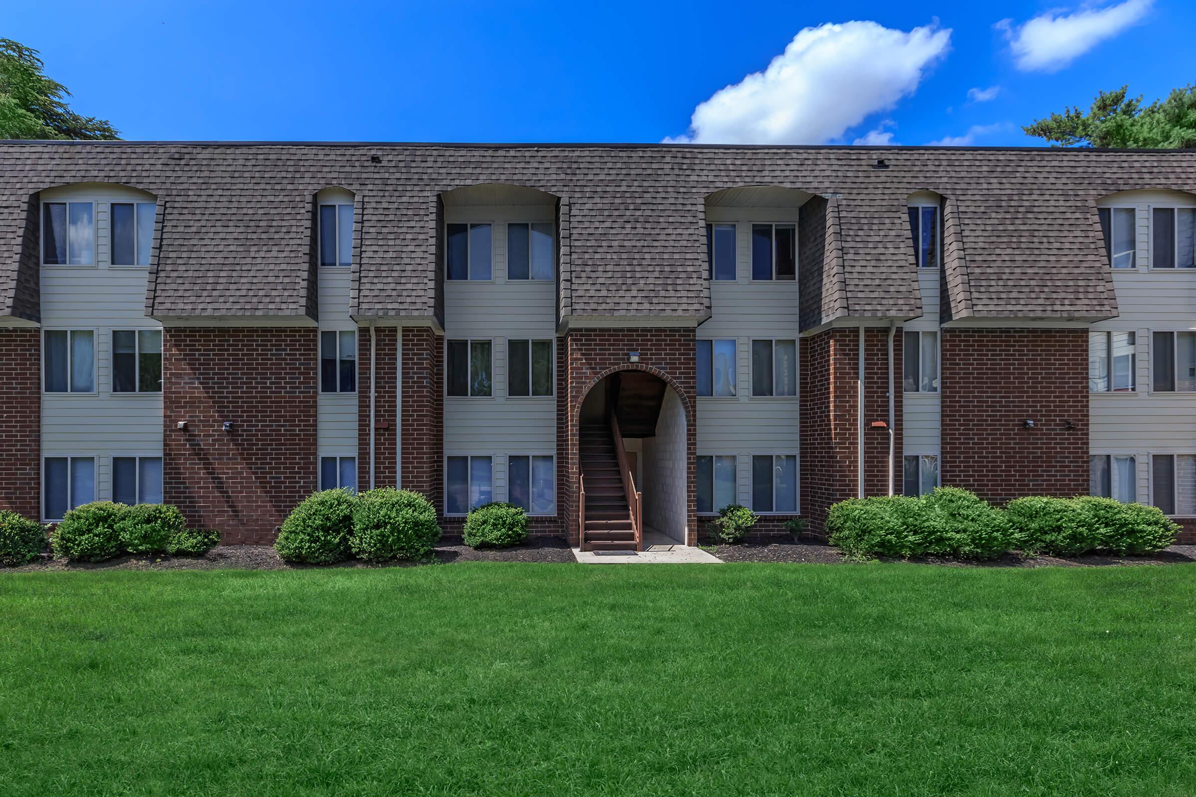 A two-story apartment building with a brick exterior and cream-colored siding. The entrance features an archway with stairs leading up. Lush green grass surrounds the building, and the sky above is bright blue with a few clouds. Neat shrubs are planted along the foundation of the building.