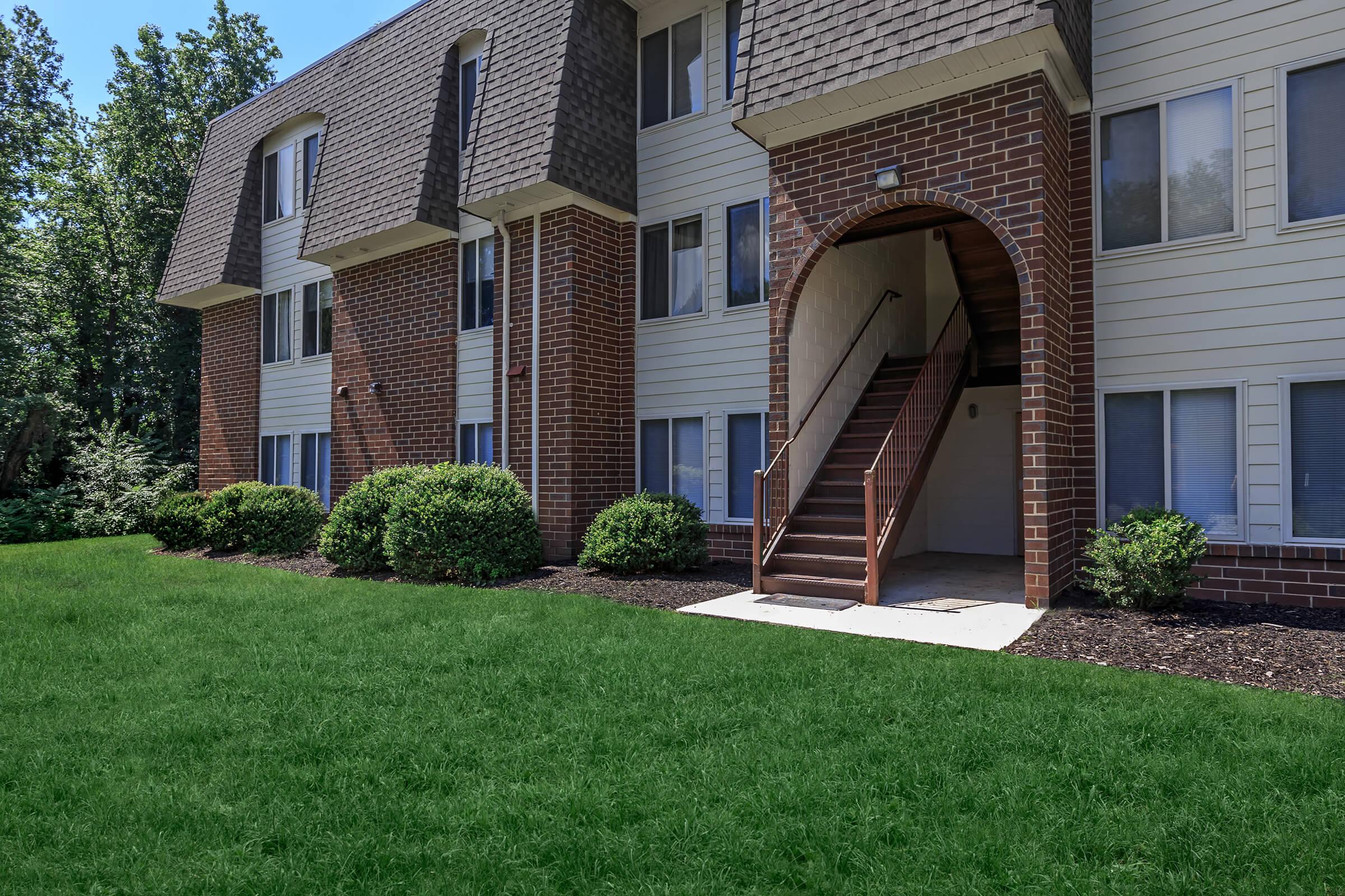 A residential apartment building featuring a brick and siding exterior. The entrance includes a set of stairs leading up to the doors, surrounded by neatly manicured green grass and small bushes. The scene is bright, with clear blue skies and a well-maintained landscape.