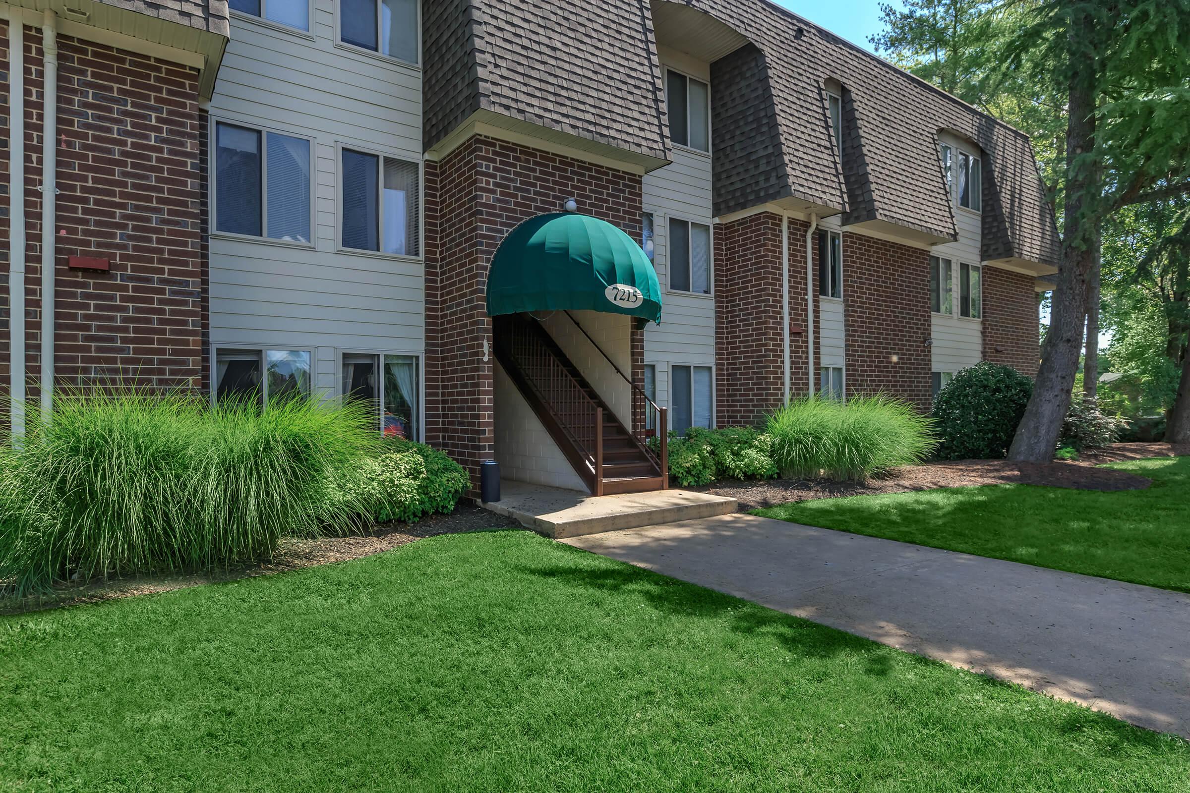 A multi-story apartment building featuring an arched green awning over the entrance, brick exterior, and large windows. The pathway leading to the entrance is lined with landscaped greenery, including grassy areas and bushes. Sunlight illuminates the scene, creating a welcoming atmosphere.