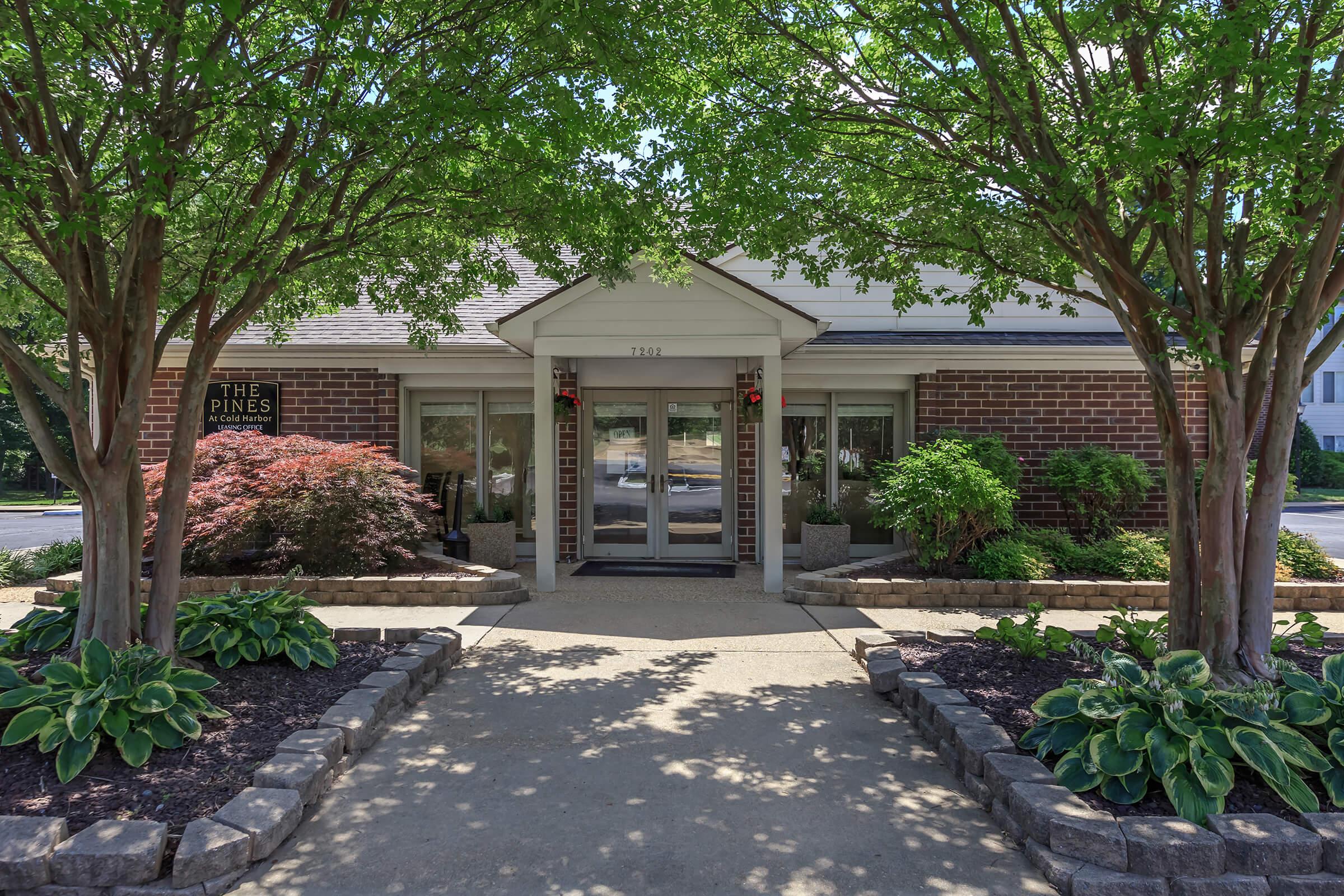 A well-maintained entrance to a building, framed by lush green trees and vibrant shrubs. The pathway is lined with decorative stones and hostas, leading to glass doors. The sign on the left reads "The Pines," indicating the name of the establishment. The setting is bright and inviting, showcasing a landscaped area.