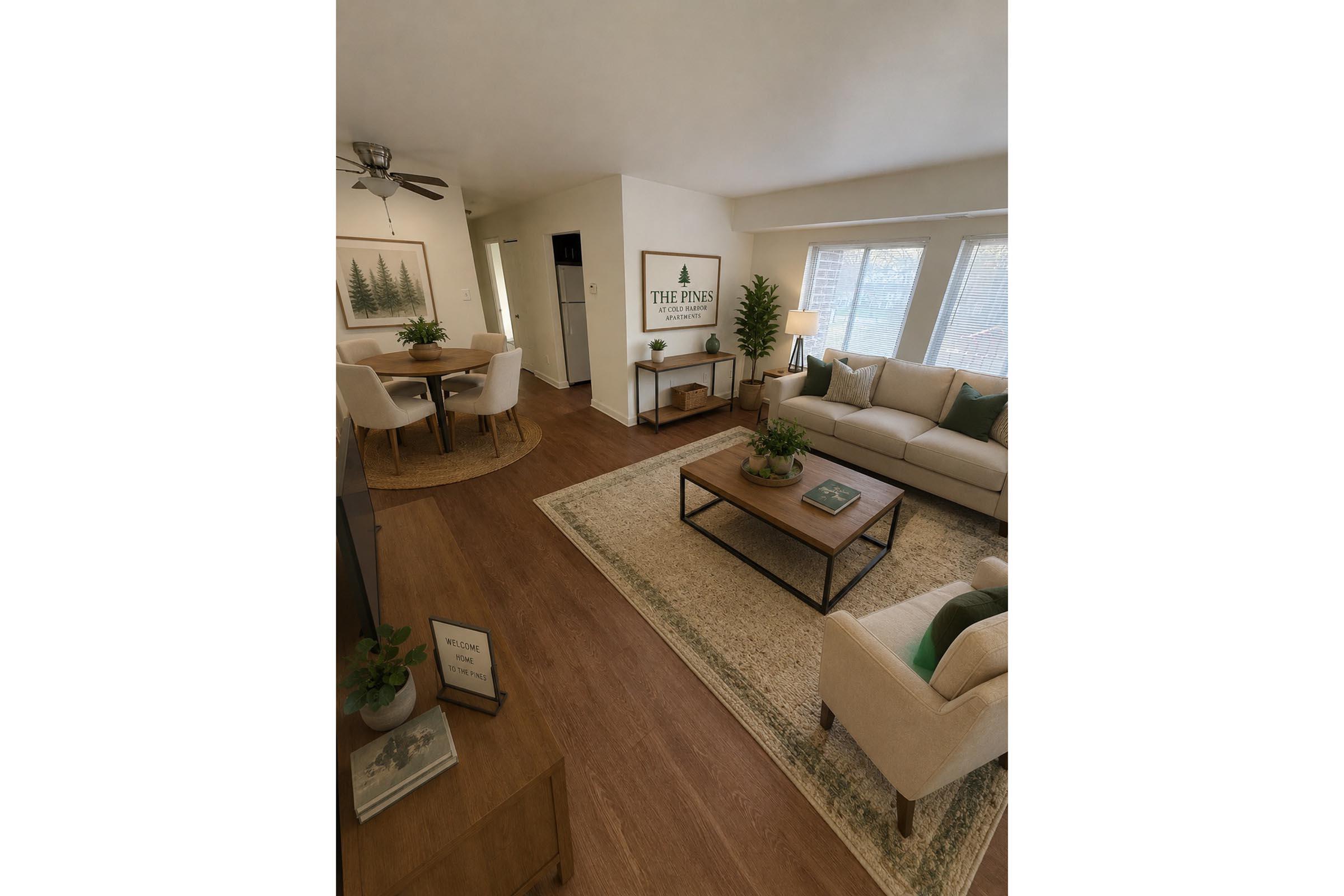 A spacious living room featuring a light-colored sectional sofa, a coffee table with decorative items, and a stylish rug. Natural light streams through large windows. In the background, there is a dining area with a round table and four chairs, and artwork on the wall showcasing trees and nature.
