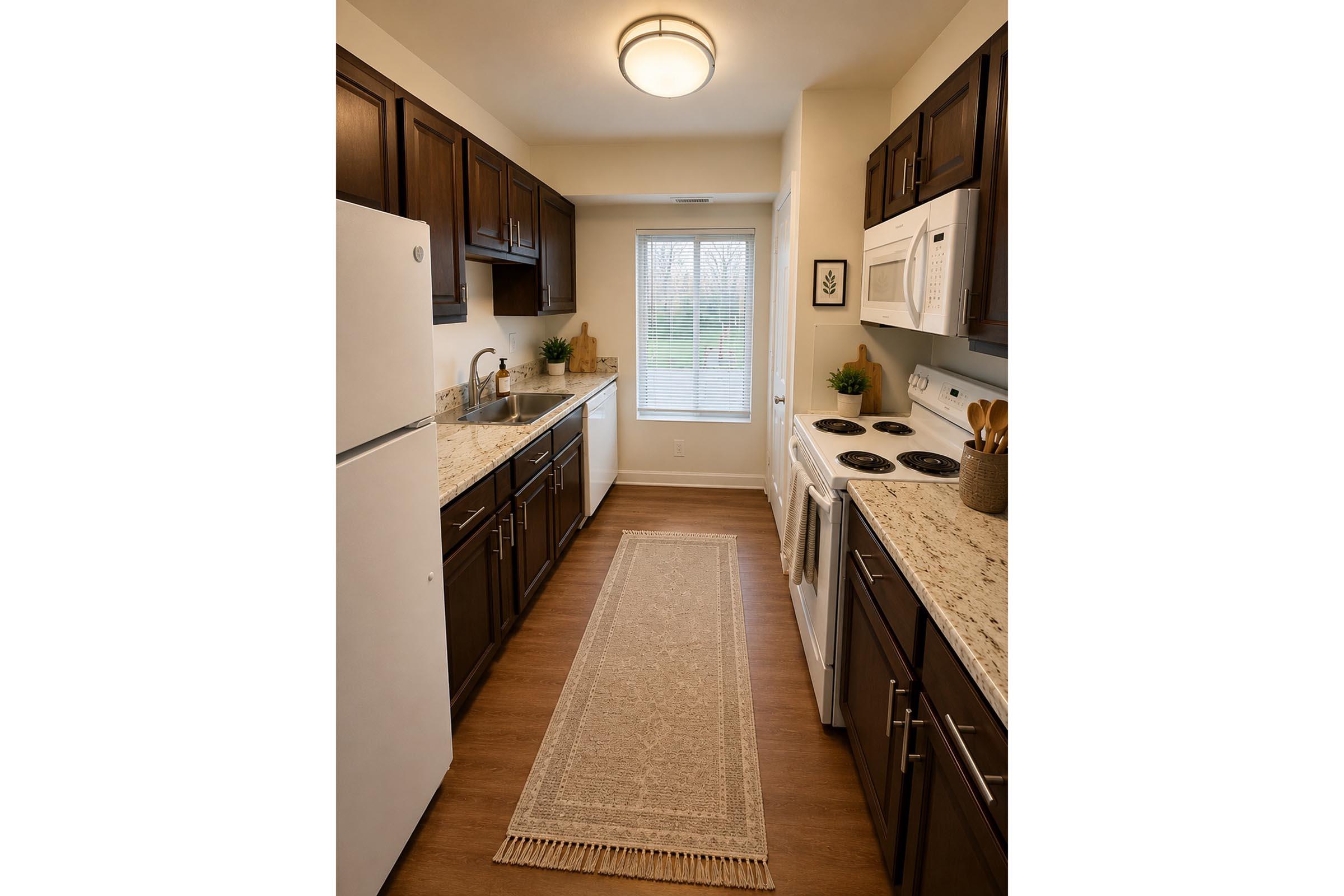 A modern kitchen featuring dark wood cabinetry, light granite countertops, a white refrigerator, and a white stove and microwave. A narrow, decorative rug runs along the floor. A window with natural light is visible, and potted plants are placed on the countertops for added decor.