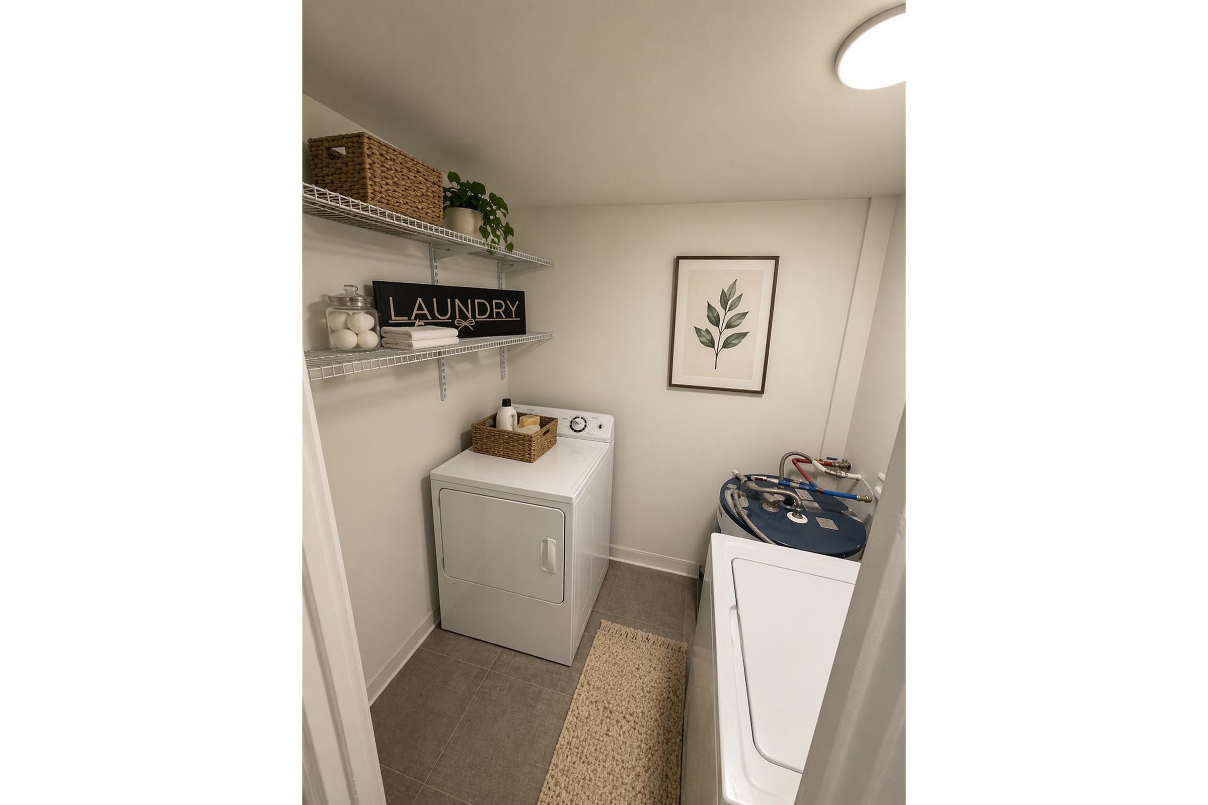 A neatly organized laundry room featuring a white washing machine and dryer, a shelf with decorative baskets, and a framed plant print on the wall. A sign reading "LAUNDRY" is prominently displayed. The space has light-colored walls and a neutral rug on the floor.