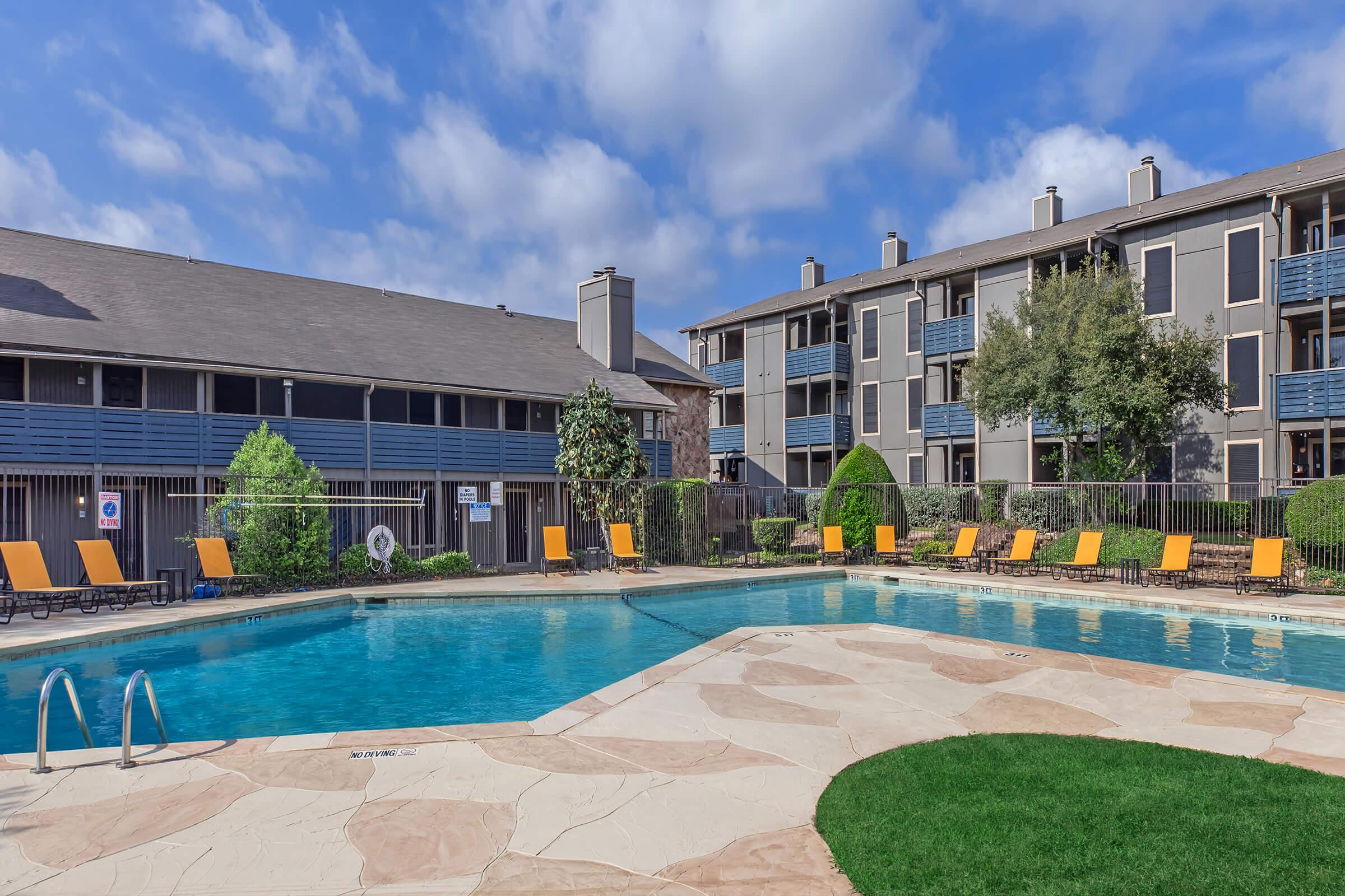 A serene swimming pool surrounded by lounge chairs, with modern apartment buildings in the background. Lush greenery and bright blue skies enhance the inviting atmosphere of this outdoor space. The pool area features a light-colored stone deck and well-maintained landscaping.