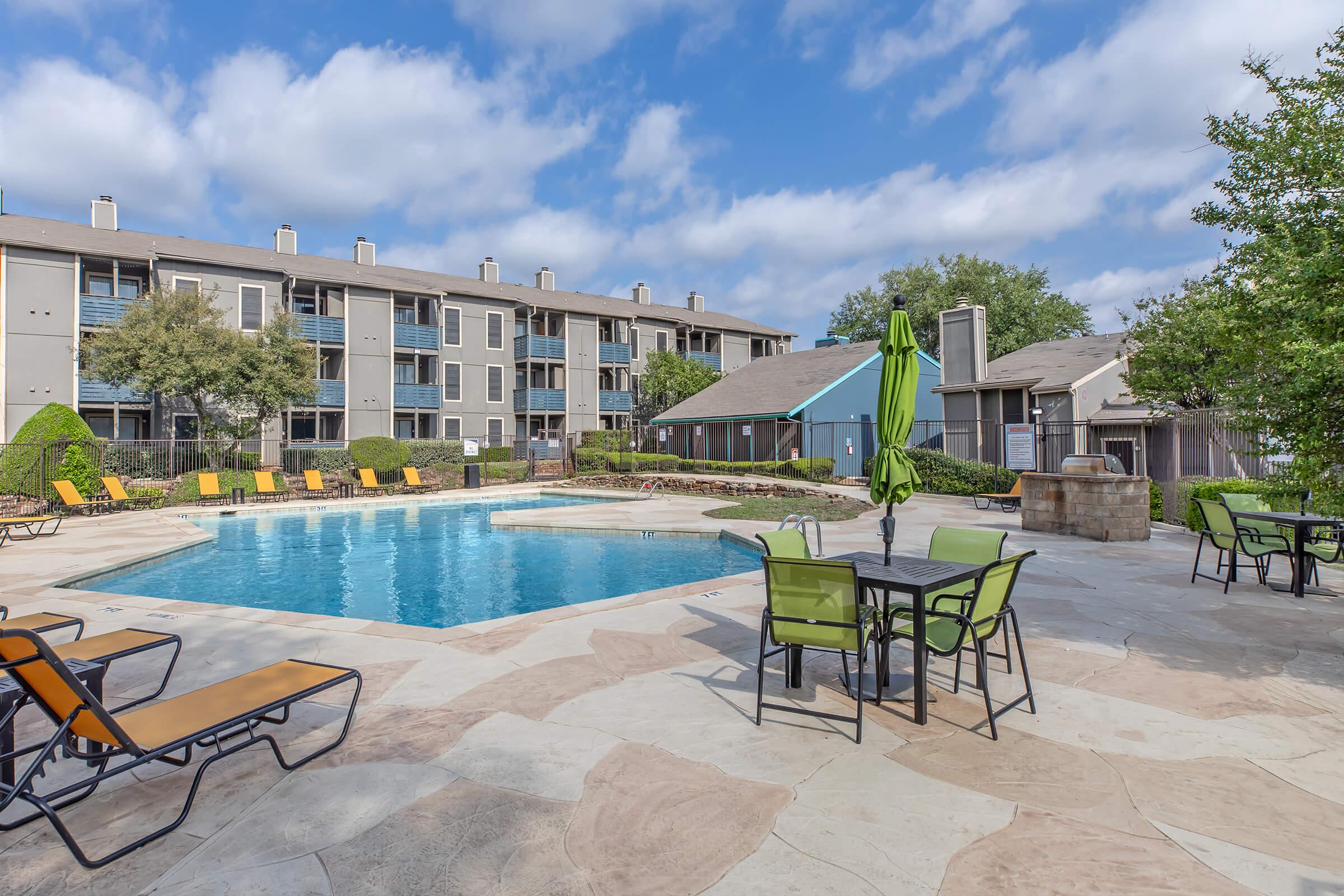 View of a well-maintained pool area featuring a clear blue swimming pool surrounded by lounge chairs and green umbrellas. There are landscaped trees and bushes, with multi-story apartment buildings in the background under a partly cloudy sky. Ideal for leisure and relaxation.