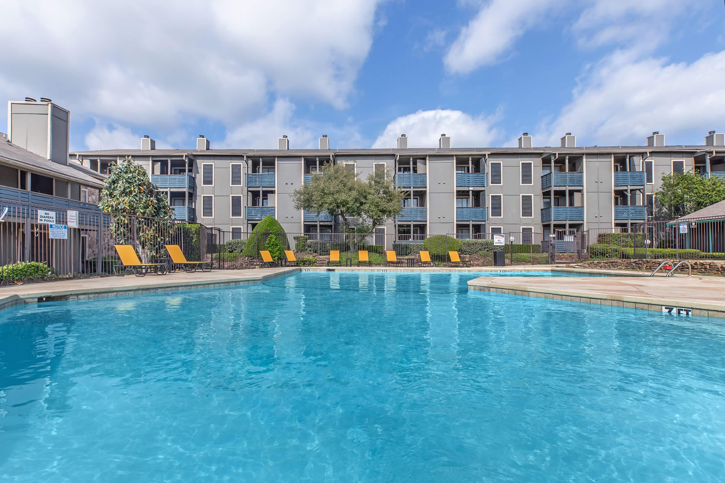 A bright, inviting swimming pool surrounded by lounge chairs in a residential area. In the background, a multi-story apartment building is visible under a partly cloudy sky. Lush greenery adds to the serene atmosphere of the setting.