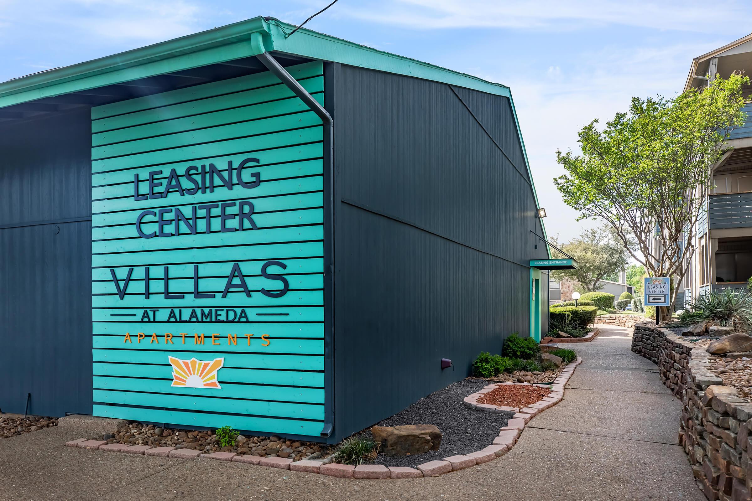 Leasing Center for Villas at Alameda Apartments, featuring a turquoise and blue exterior with bold signage. A landscaped walkway with decorative rocks leads to the entrance, surrounded by greenery and a clear sky.