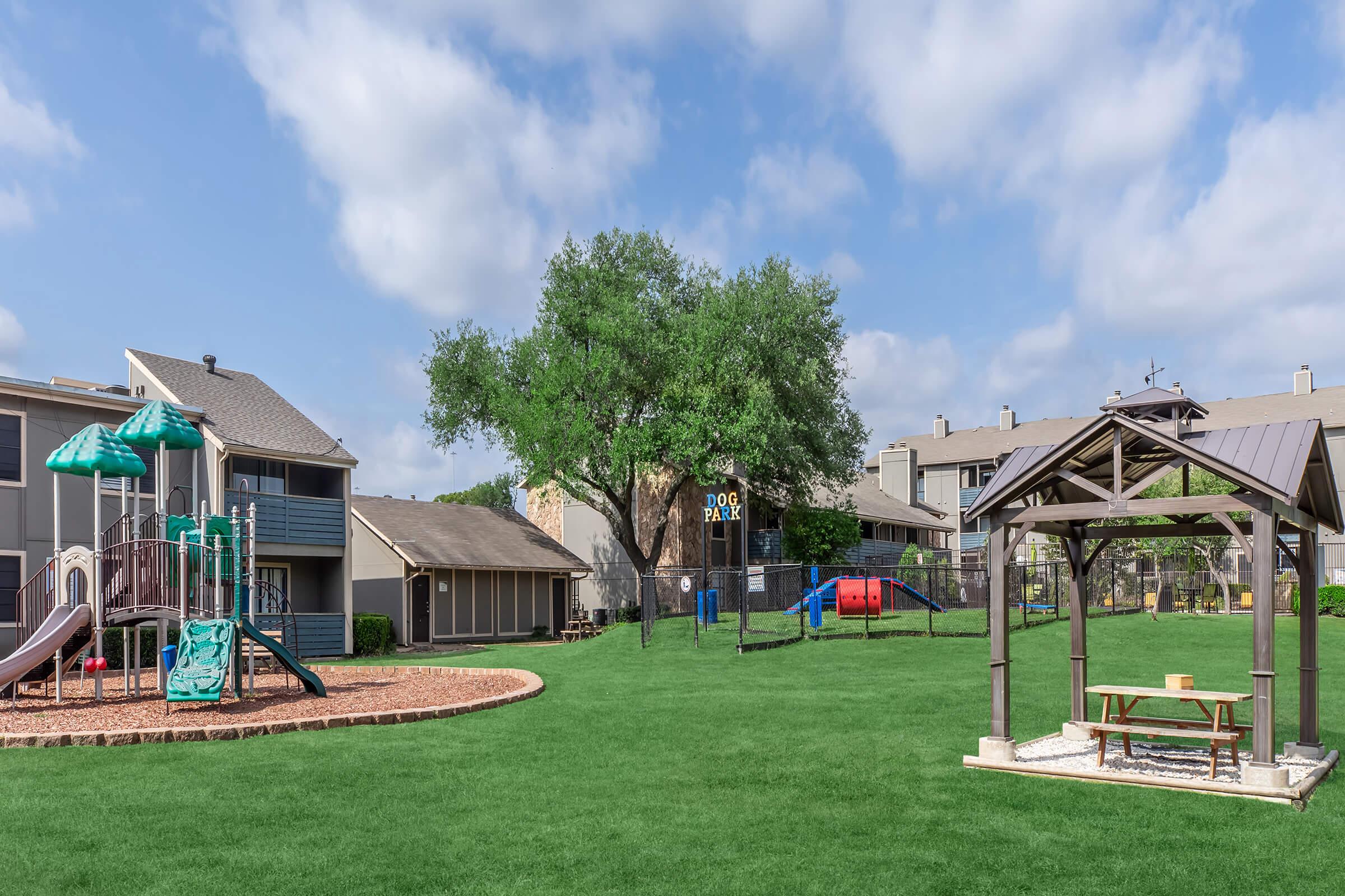 A spacious backyard area featuring a playground with slides and swings, a gazebo with a table, and a well-maintained lawn. In the background, there are apartment buildings and a dog park with various play equipment and a fence. The sky is partly cloudy, creating a bright and inviting atmosphere.