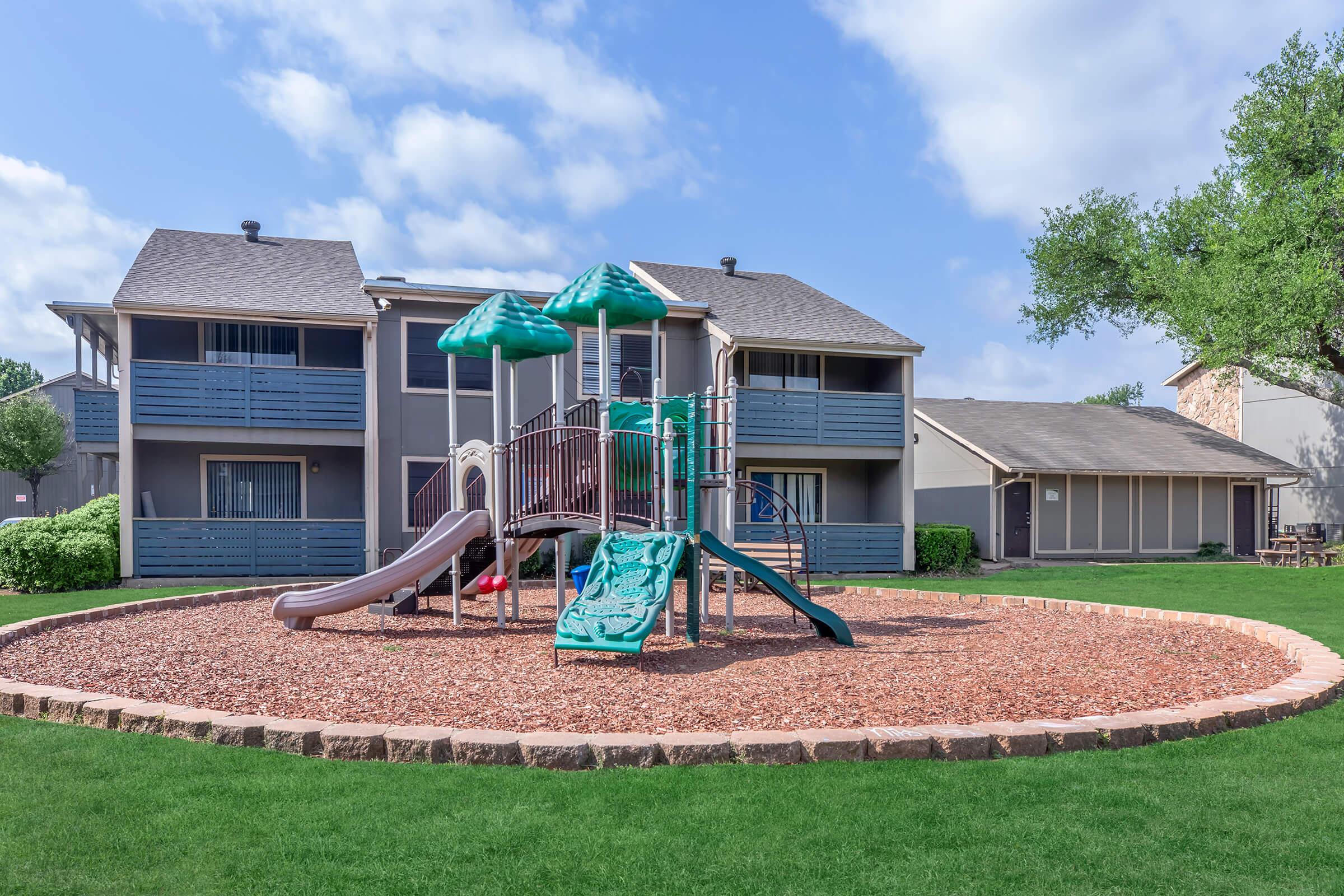 Playground with two slides and climbing equipment surrounded by a circular area of mulch. In the background, there are two gray apartment buildings with balconies and windows. The sky is partly cloudy, and the area is well-maintained with green grass and shrubs.