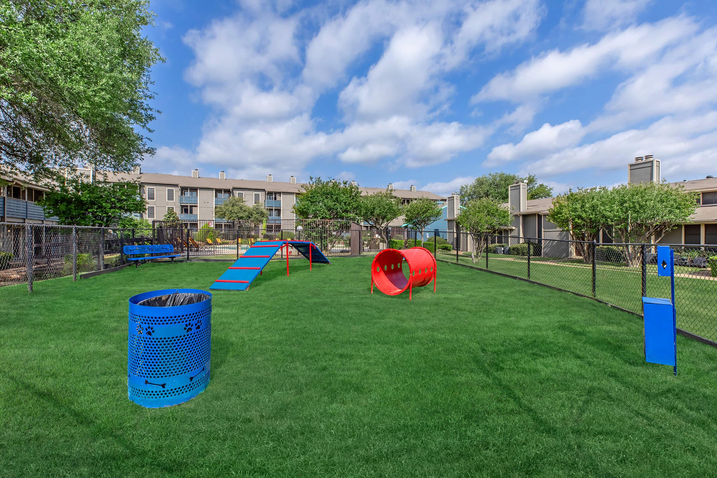 A spacious outdoor dog park featuring green grass, blue agility equipment, and a red tunnel. The park is surrounded by a fence and several trees. In the background, there are residential buildings under a partly cloudy sky.
