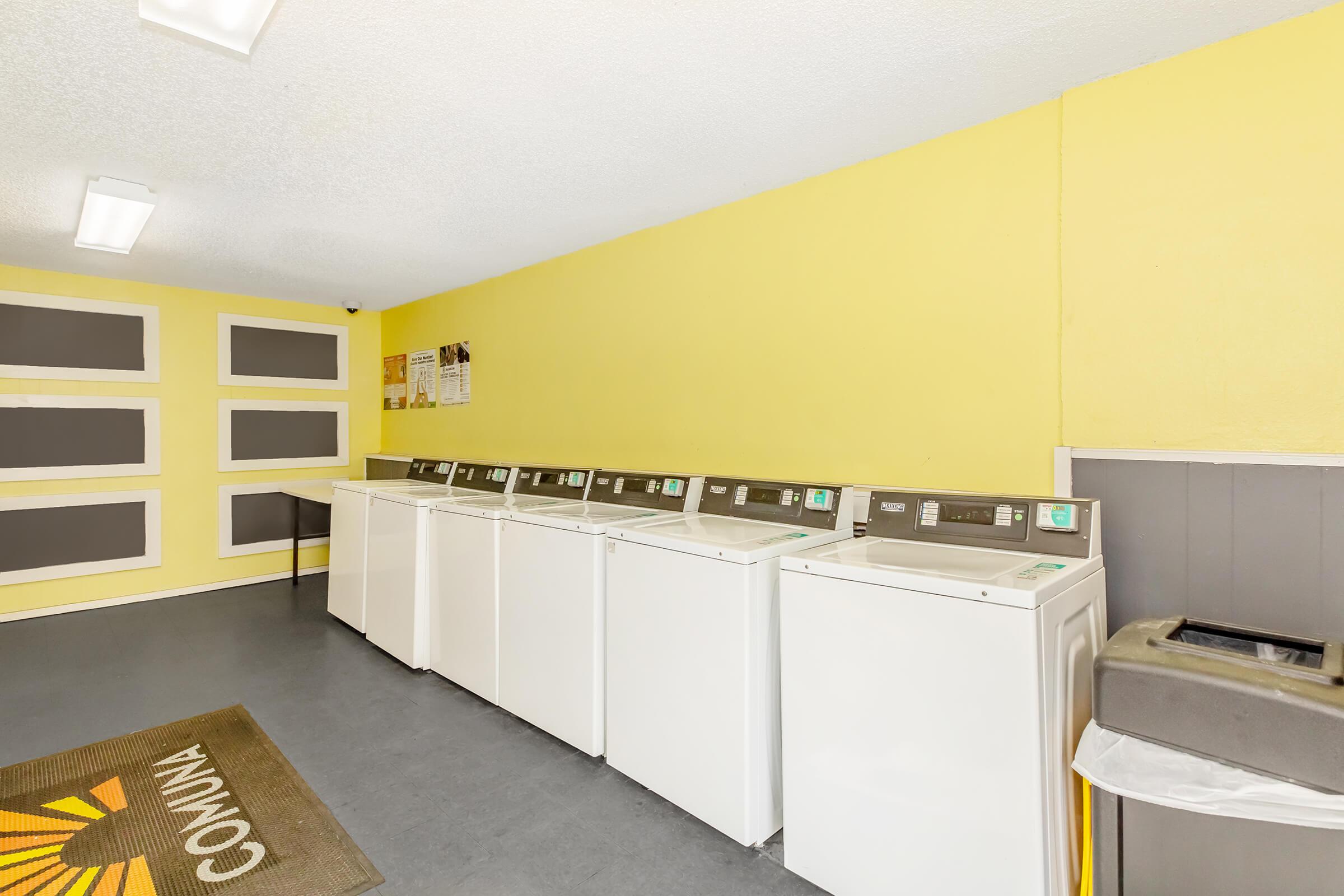 A bright, clean laundry room featuring multiple white washing machines aligned against a yellow wall. There's a welcome mat at the entrance and a trash bin nearby. The room is well-lit with ceiling lights and has bulletin boards displaying information.